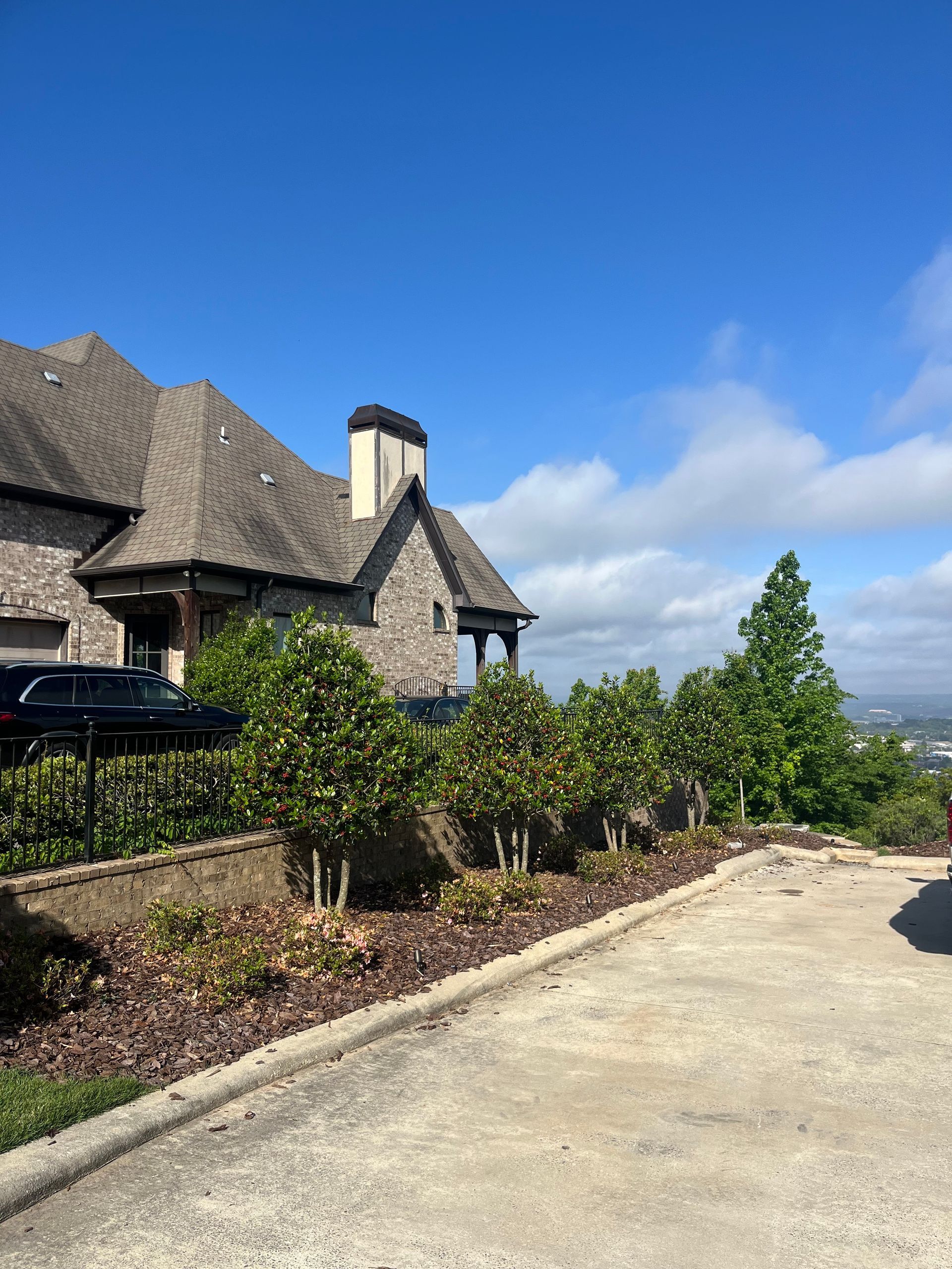 Stone house with steep roof beside a gravel drive, landscaped shrubs, and a blue sky with clouds.