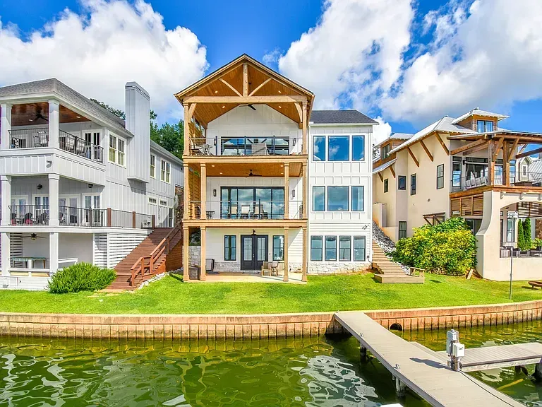 Waterfront row of modern homes with balconies, green lawns, and docks along a canal under a blue sky.