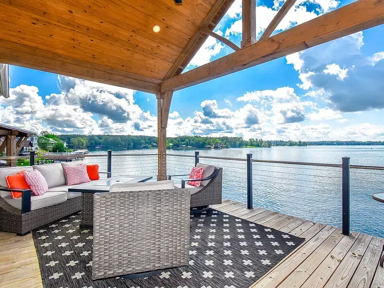 Lakeside deck with wicker seating, patterned rug, and wooden roof overlooking calm water and clouds