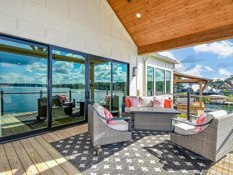 Covered patio with wicker seating, patterned rug, and large glass doors overlooking the water and sky.