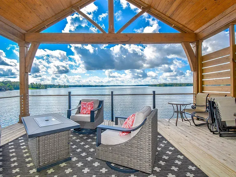 Covered lakeside deck with wicker chairs, table, and wheelchair, overlooking calm water under a blue sky.