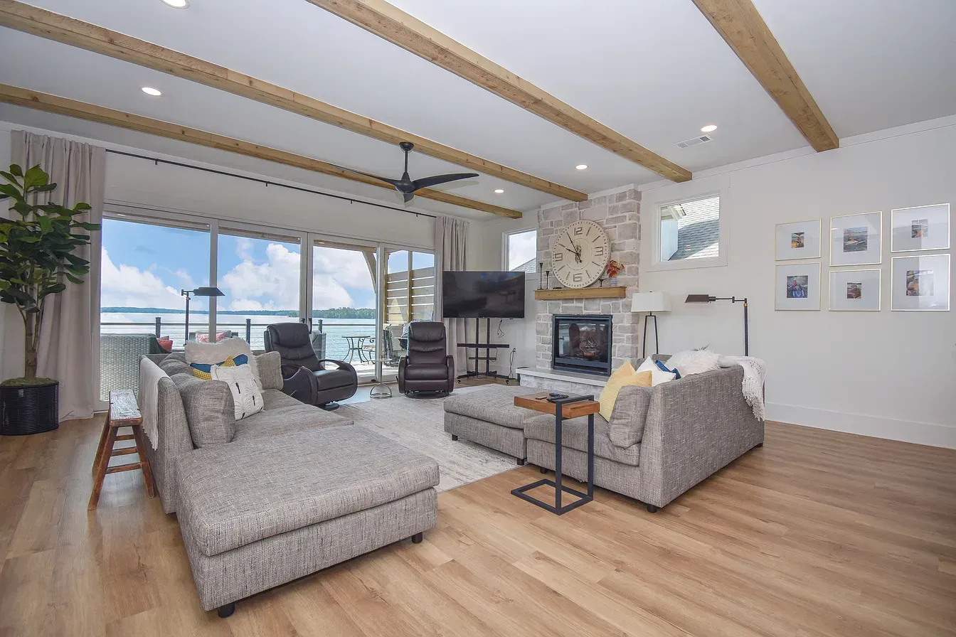 Bright living room with gray sectional, fireplace, TV, wood beams, and large bay windows overlooking water