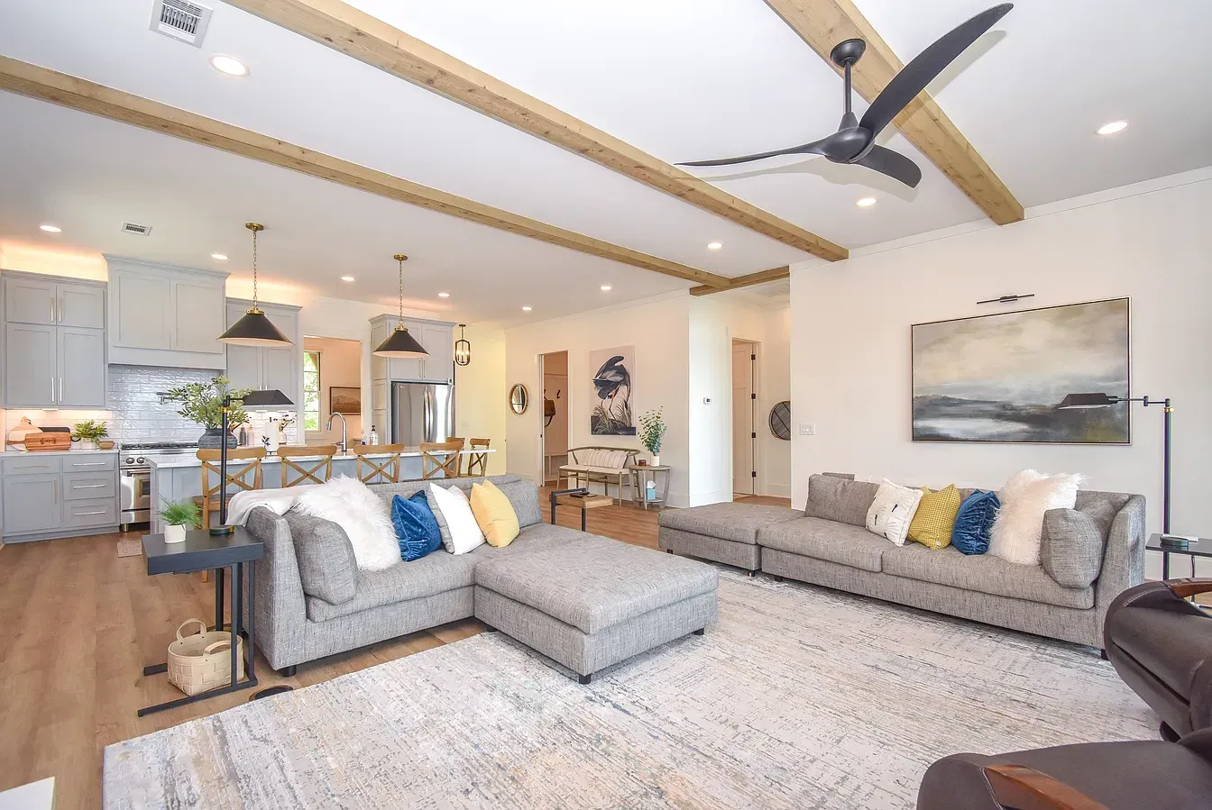 Bright, open living room with gray sofas, vaulted ceiling, ceiling fan, and kitchen in the background