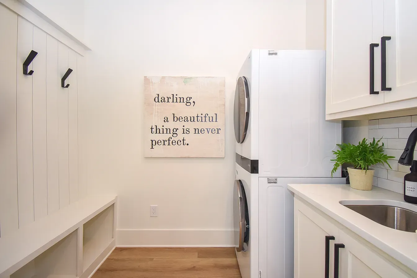 Bright laundry room with white cabinets, washer and dryer, wood floor, and a wall sign.