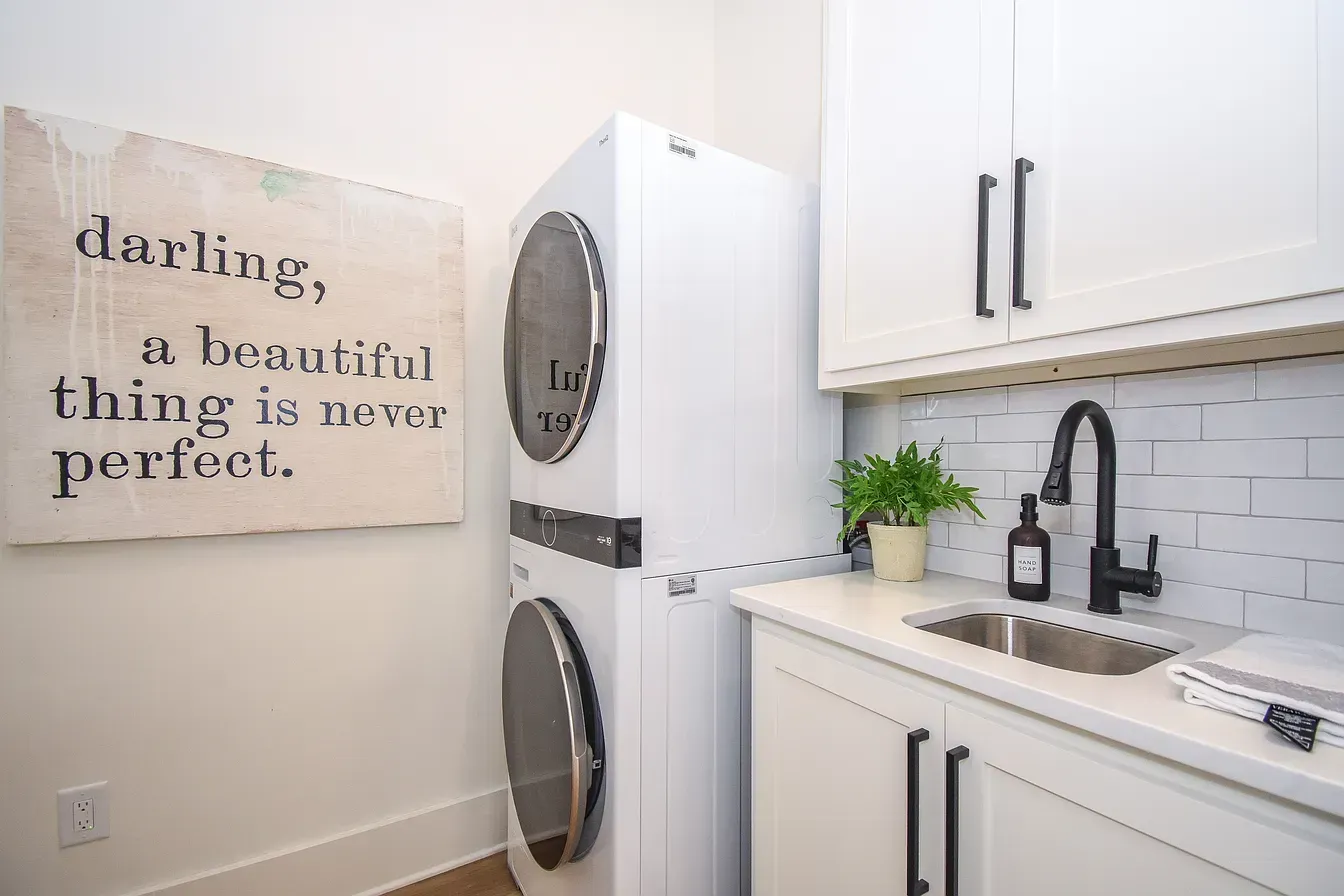 Laundry room with white cabinets, stacked washer and dryer, sink, and wall sign reading “a beautiful thing is never perfect.”