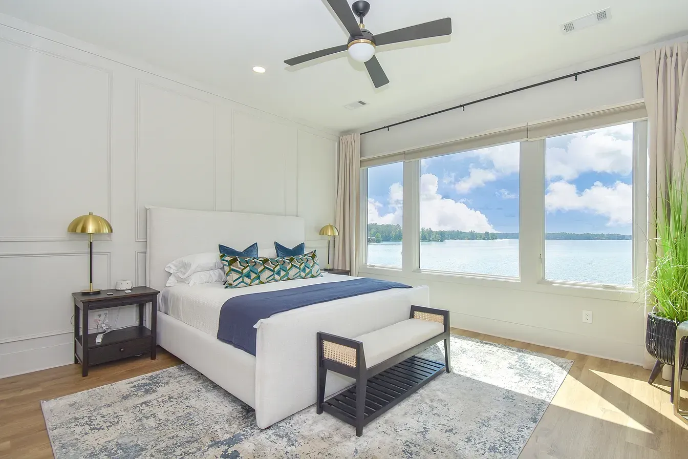 Bright bedroom with a white bed, blue accents, and large windows overlooking the water