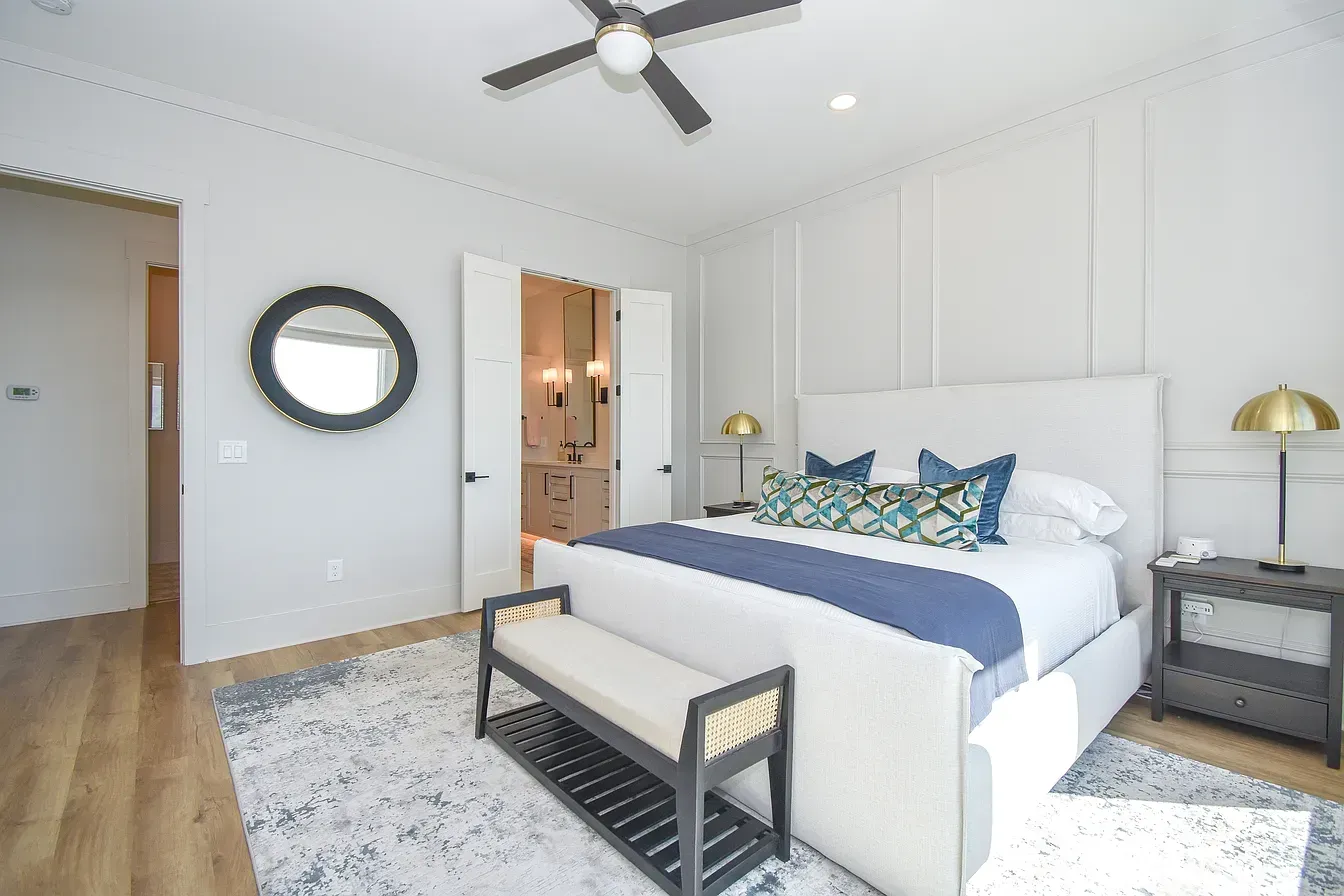 Bright bedroom with a white bed, gray rug, wooden bench, ceiling fan, and round wall mirror.