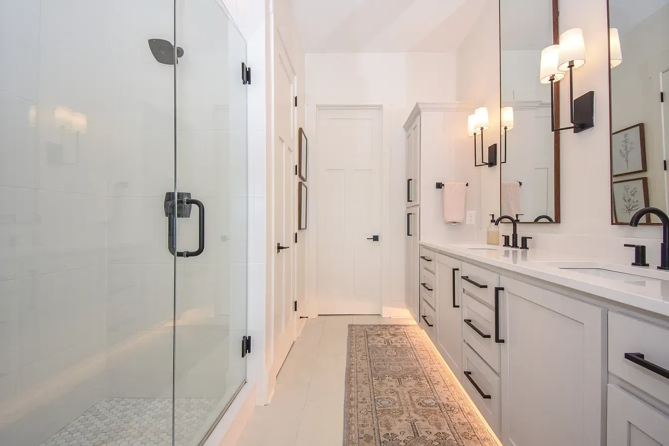 Bright white bathroom with glass shower, double vanity, wall sconces, and a gray runner rug.