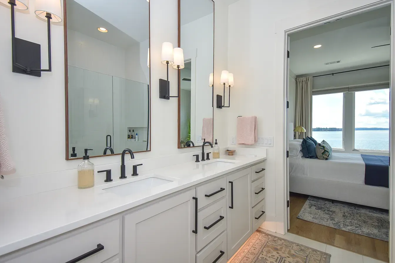 Modern bathroom with double vanity, wall sconces, and an open doorway to a bright bedroom with ocean view