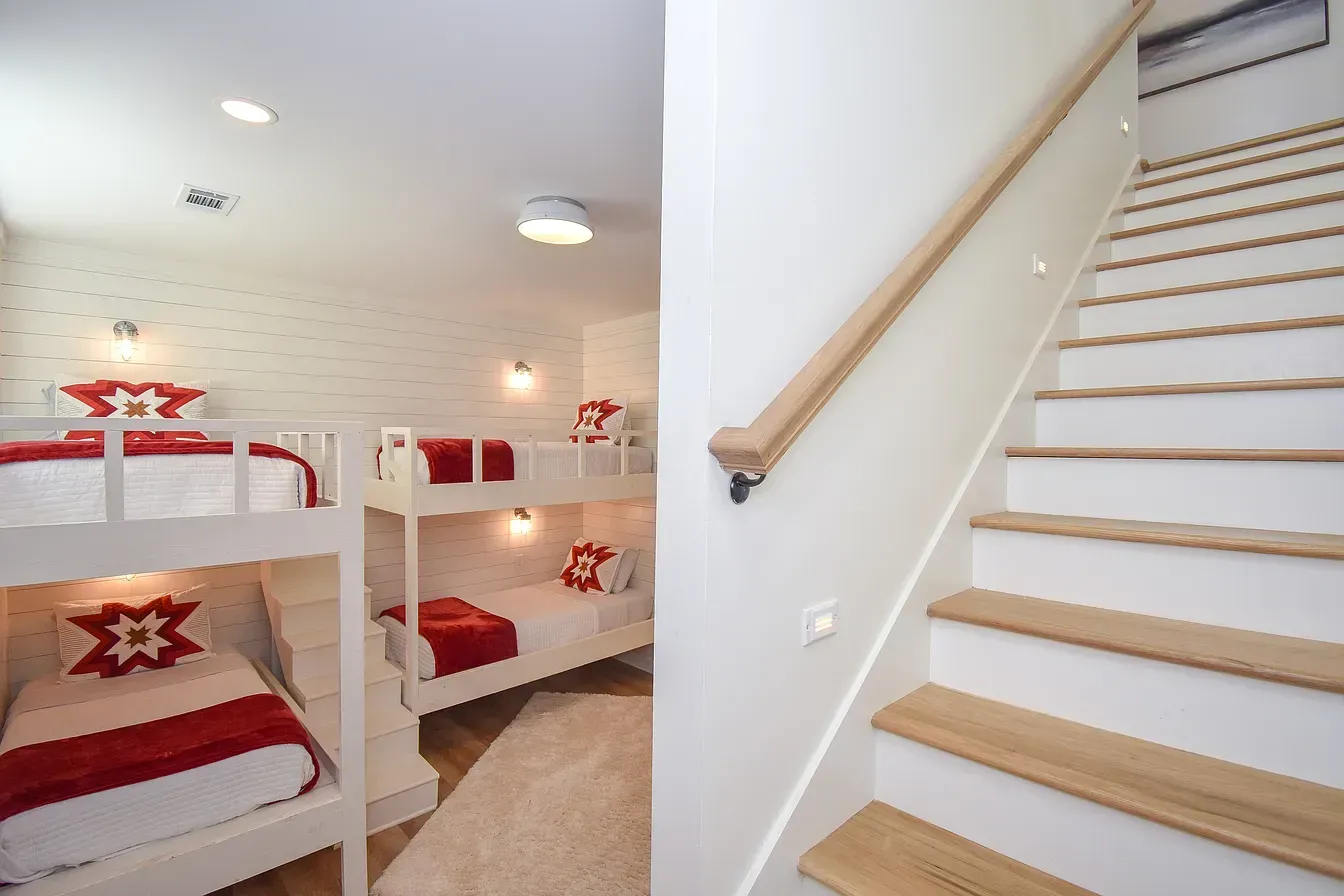 White bunk-bed dorm room beside a stairway with light wood railing and red bedding accents