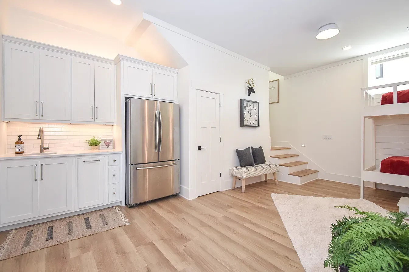 Bright open kitchen and living area with white cabinets, stainless fridge, stairs, and a bunk bed nook.
