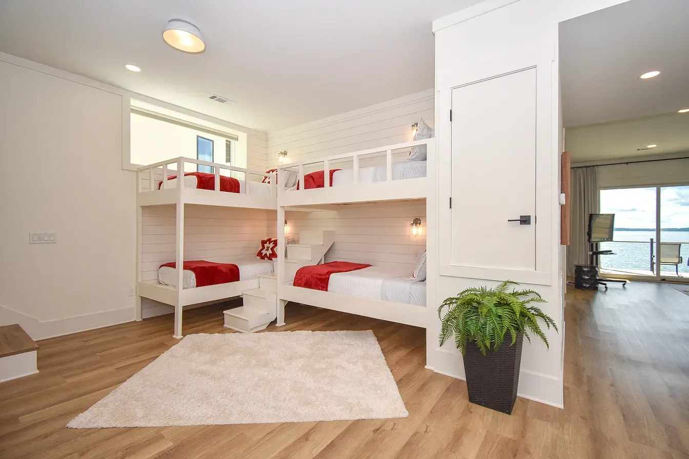 Bright shared bedroom with white bunk beds, red bedding, wood floors, and a potted plant near a doorway.