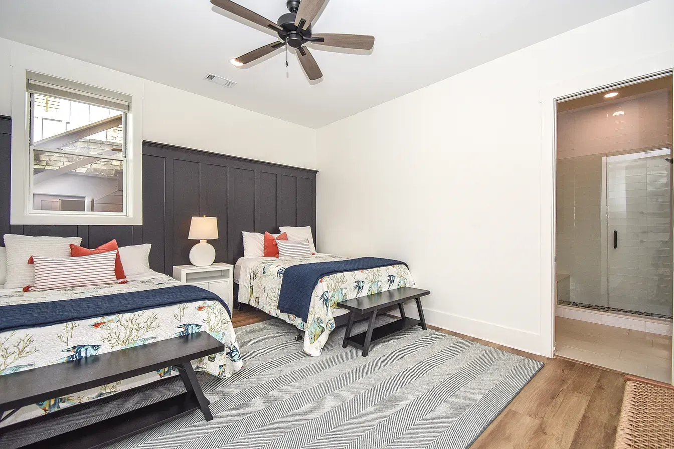 Bright bedroom with two twin beds, dark headboard, ceiling fan, patterned rug, and adjoining bathroom visible