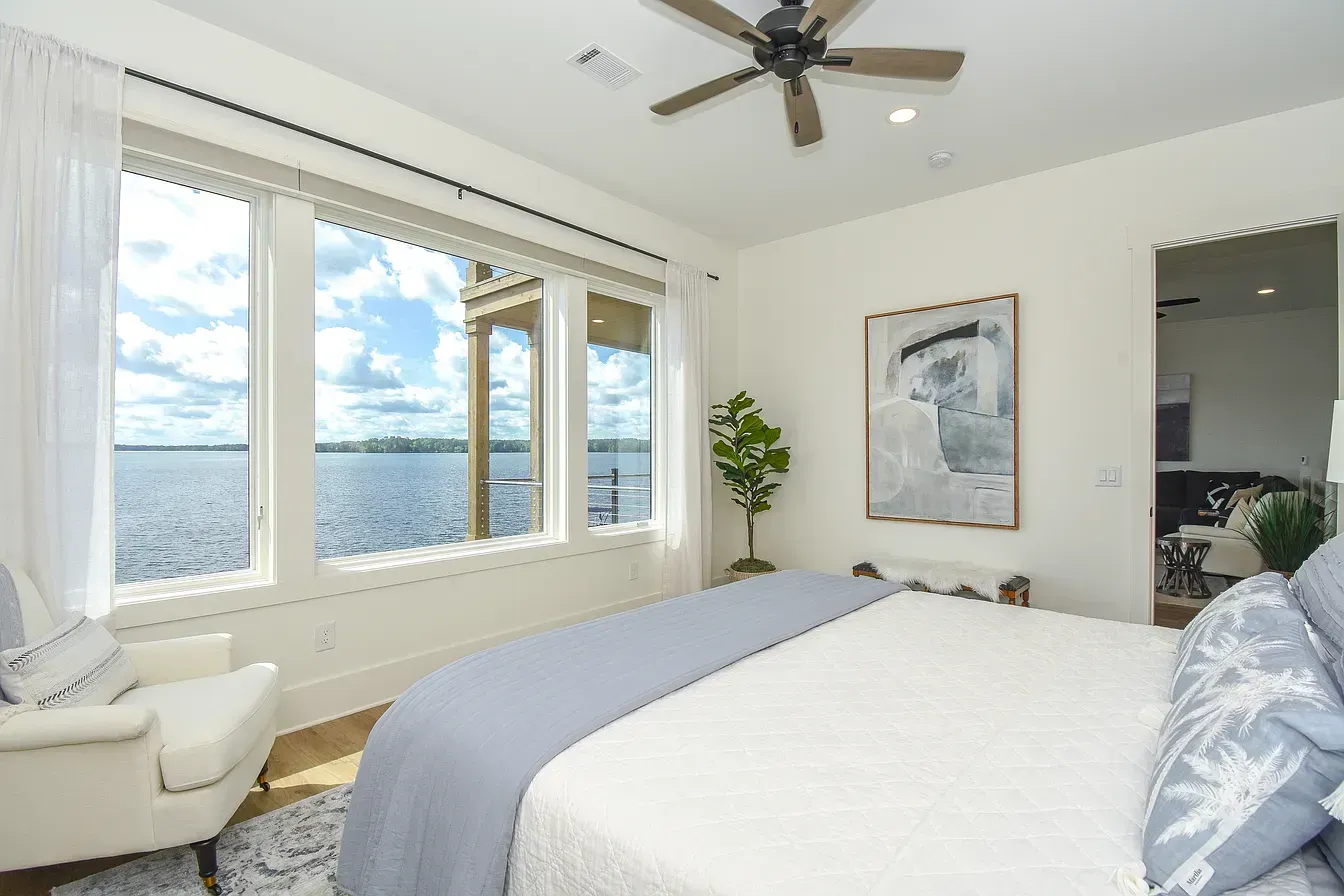 Bright bedroom with large windows overlooking water, white chair, bed, and ceiling fan