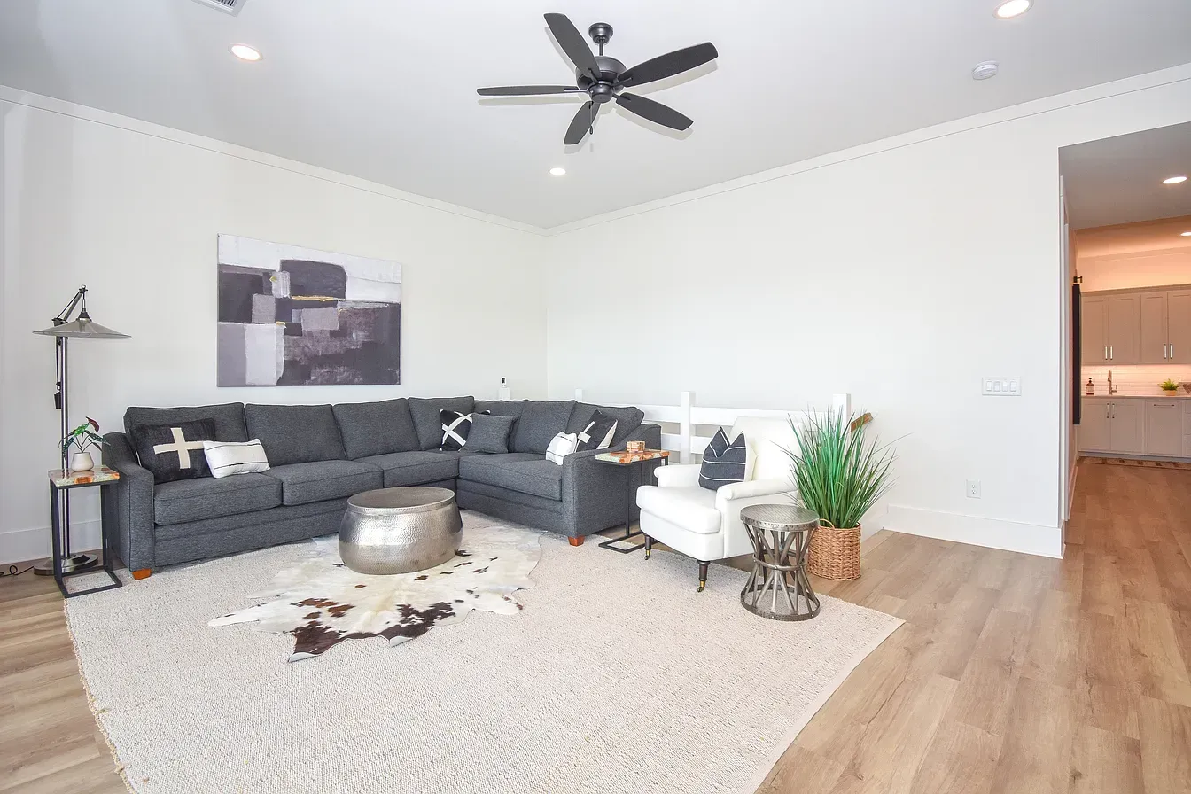 Bright living room with gray sectional, white rug, wood floor, ceiling fan, and potted plant