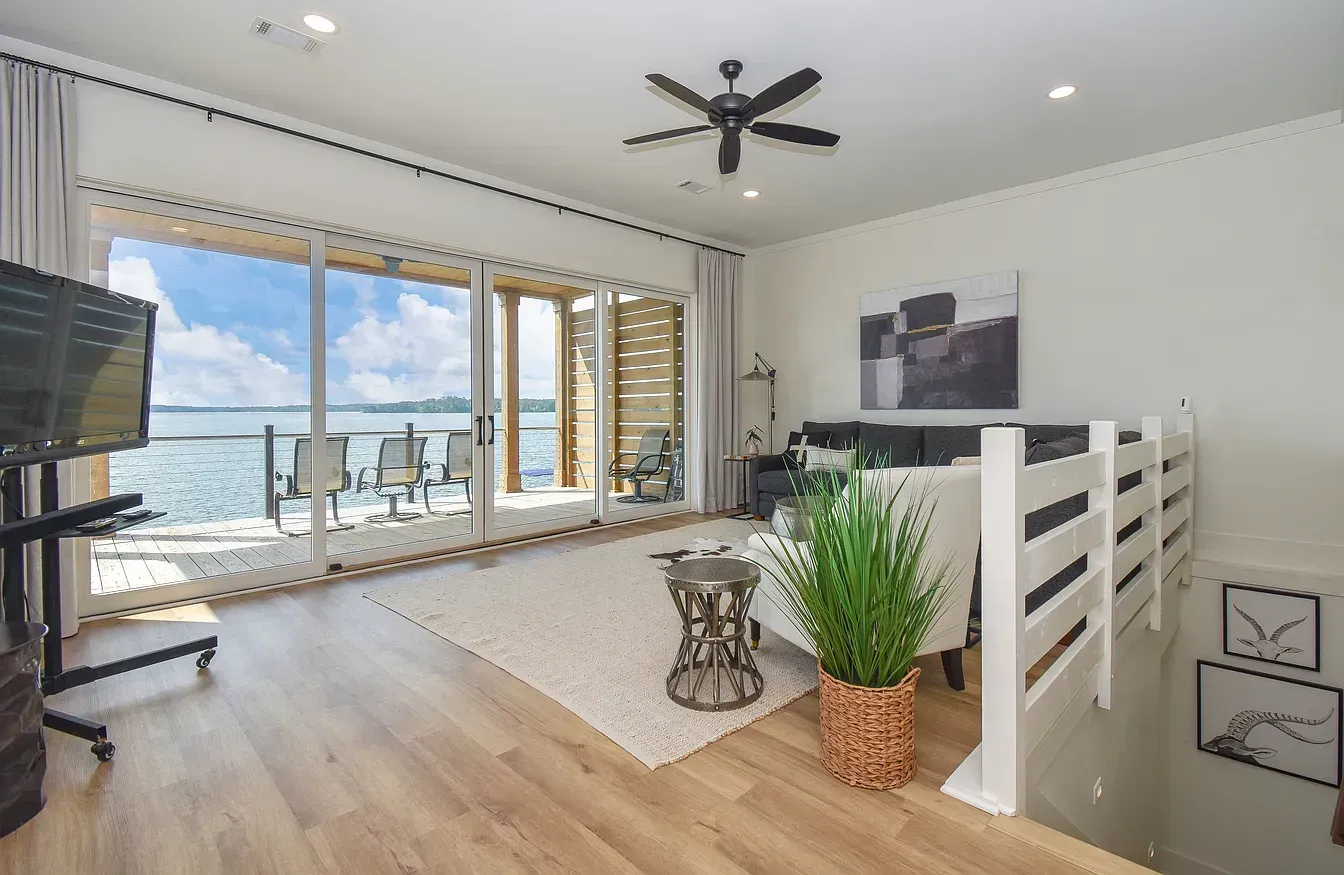 Bright modern living room with TV, wood floors, and large windows overlooking a patio and water view