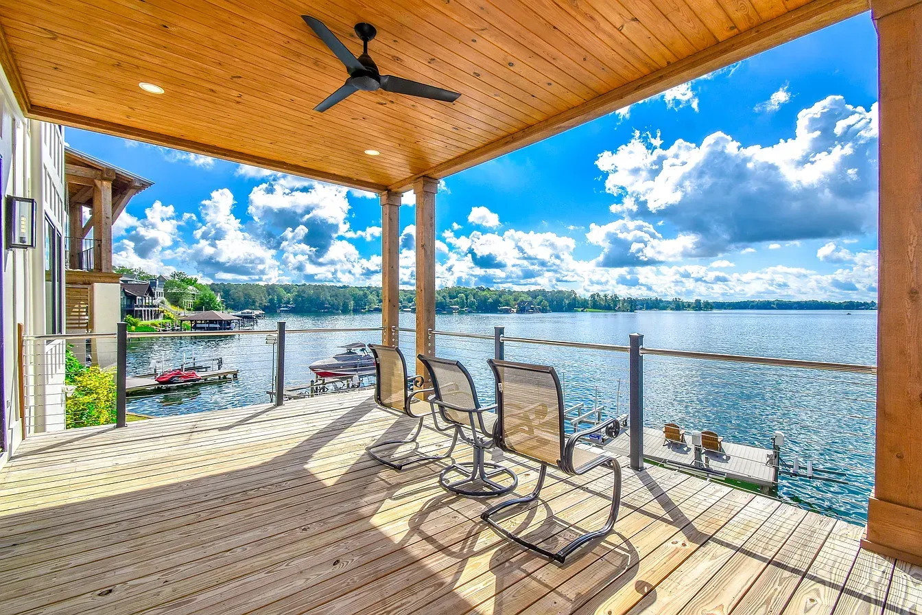 Wooden lakeside deck with chairs, ceiling fan, and a bright view of calm water and blue sky