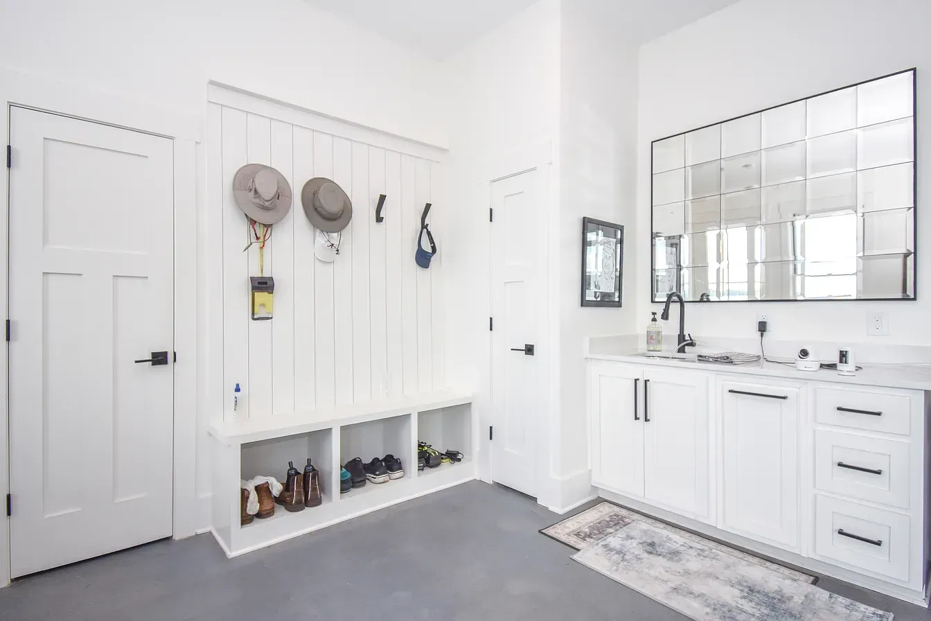 Bright white mudroom with built-in bench, hooks, storage cubbies, mirror, and sink vanity.