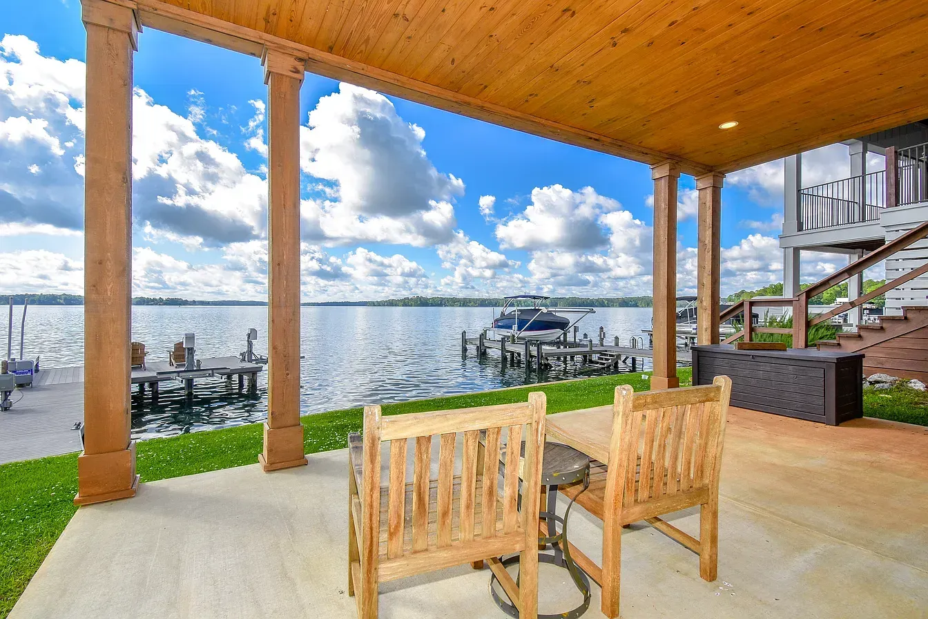 Covered lakeside deck with wooden chairs, dock, and calm water under a cloudy blue sky