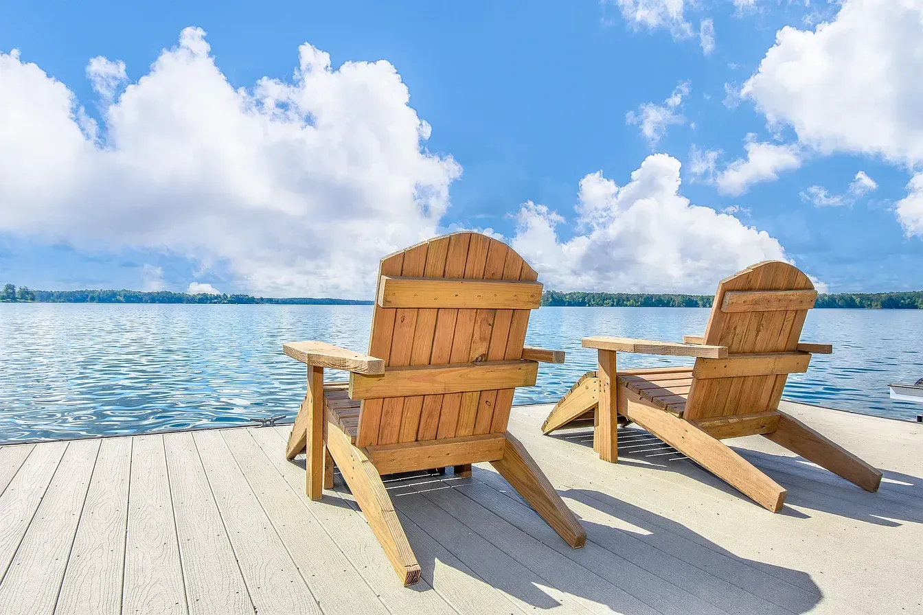Two wooden Adirondack chairs facing a calm blue lake under a bright sky with clouds