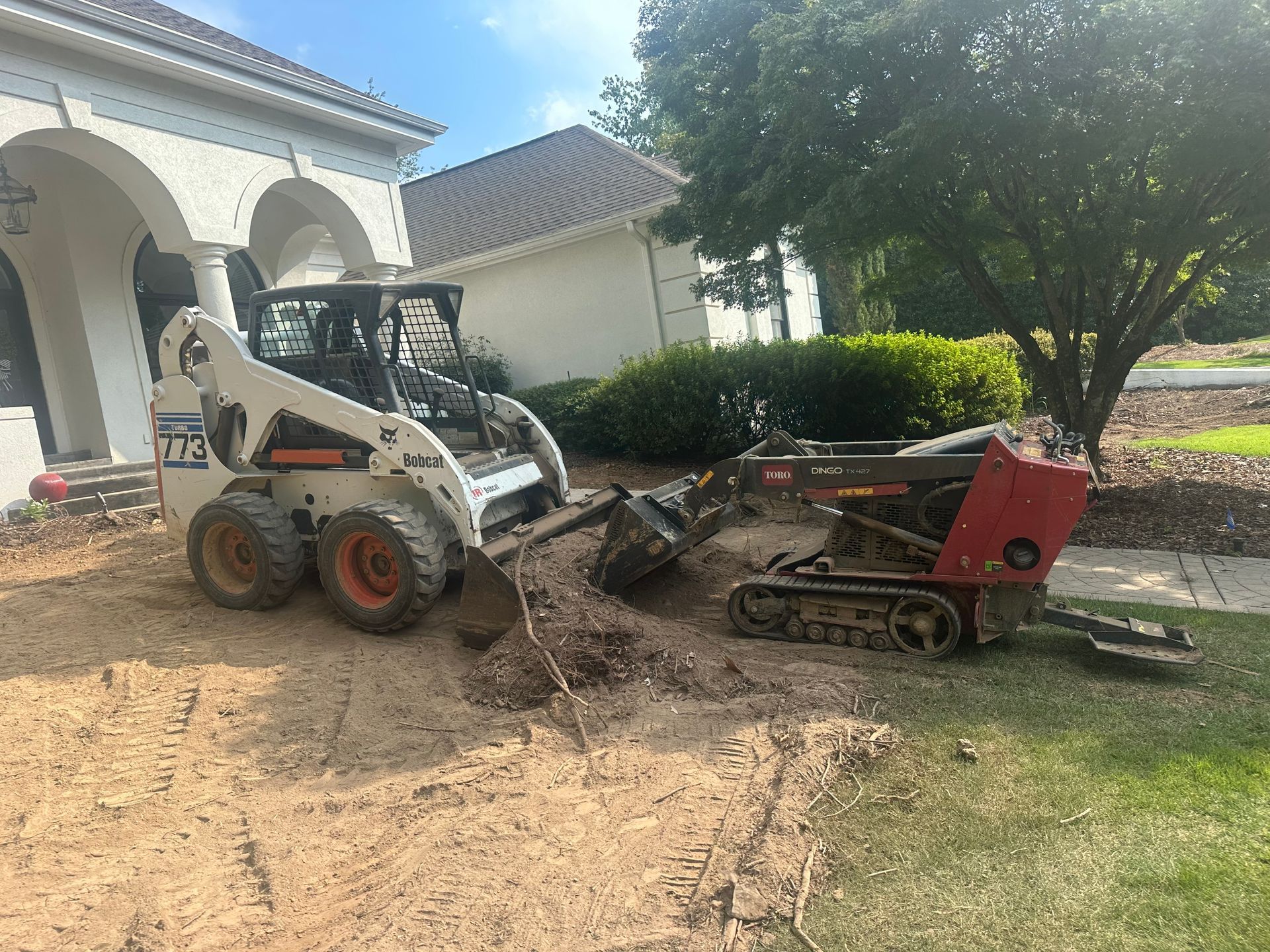 White skid steer and red tracked machine working on dirt beside a house and tree