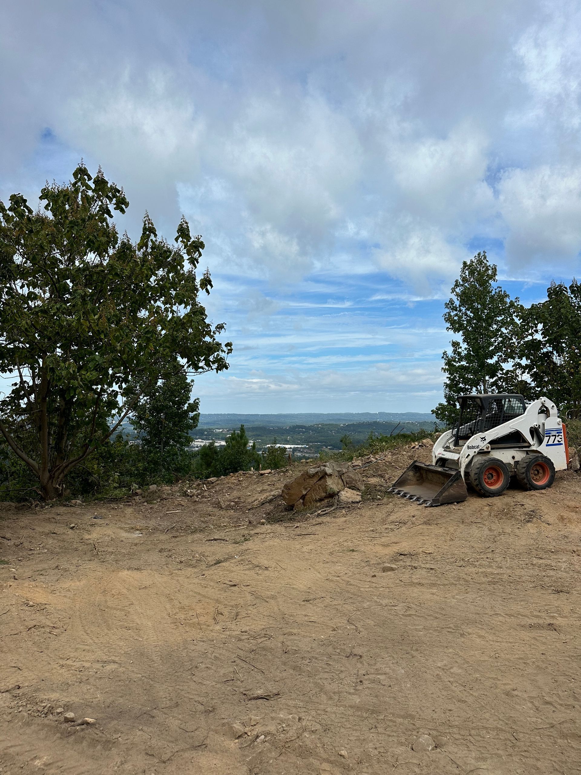 Bobcat skid-steer on a dirt overlook beside trees, with a cloudy sky and distant valley view.