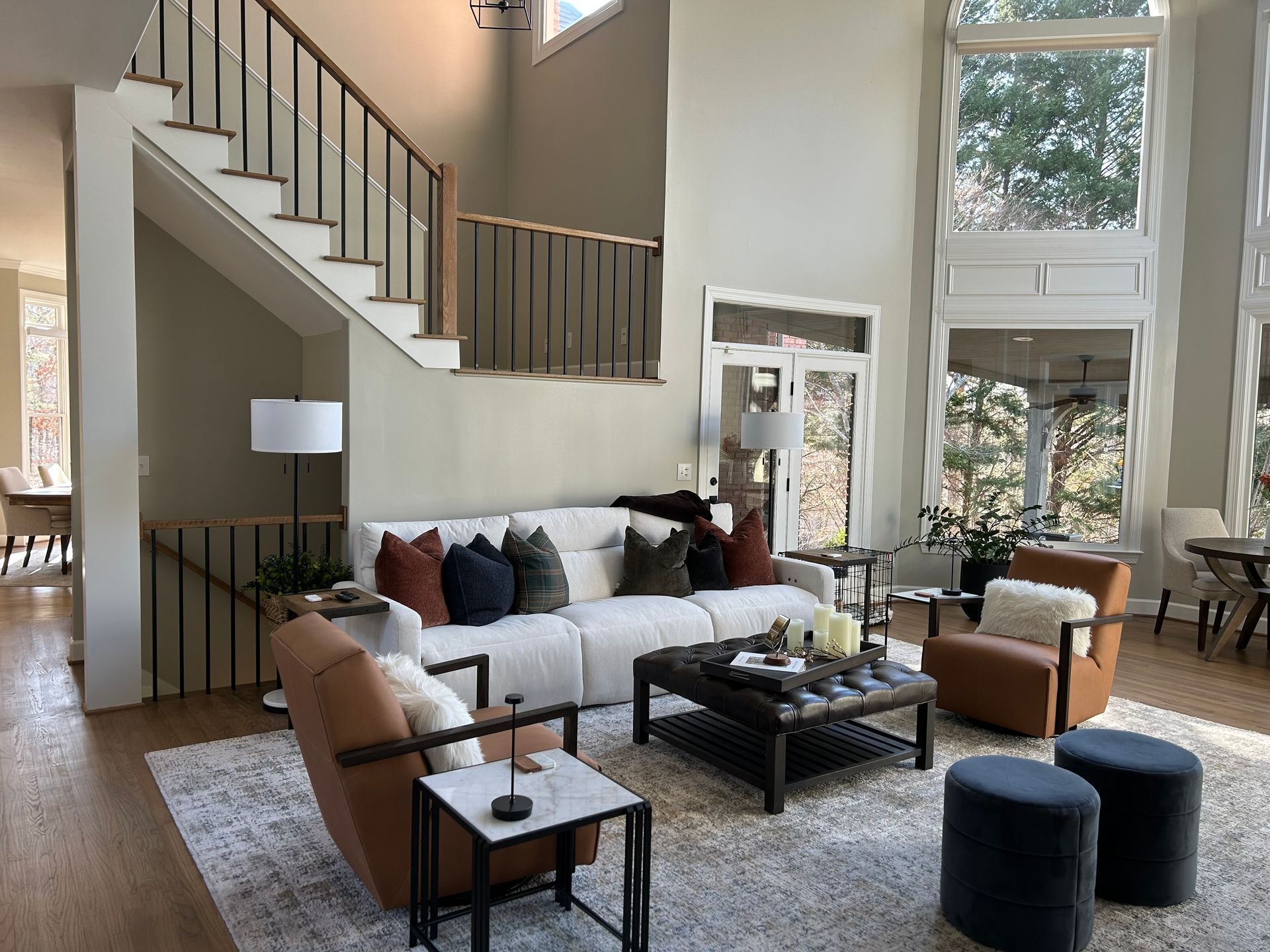 Modern living room with white sofa, tan chairs, staircase, and large windows overlooking trees