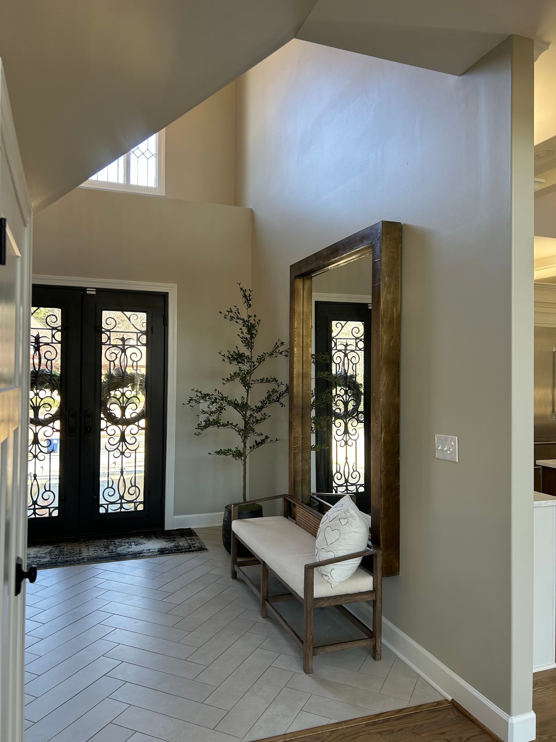 Bright foyer with tall mirror, bench, black front doors, and a decorative tree