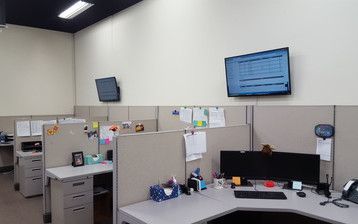 A cubicle in an office with a computer desk and a flat screen tv on the wall.