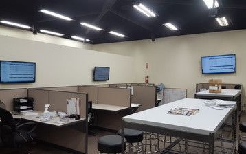 A room with cubicles , tables , chairs and televisions.