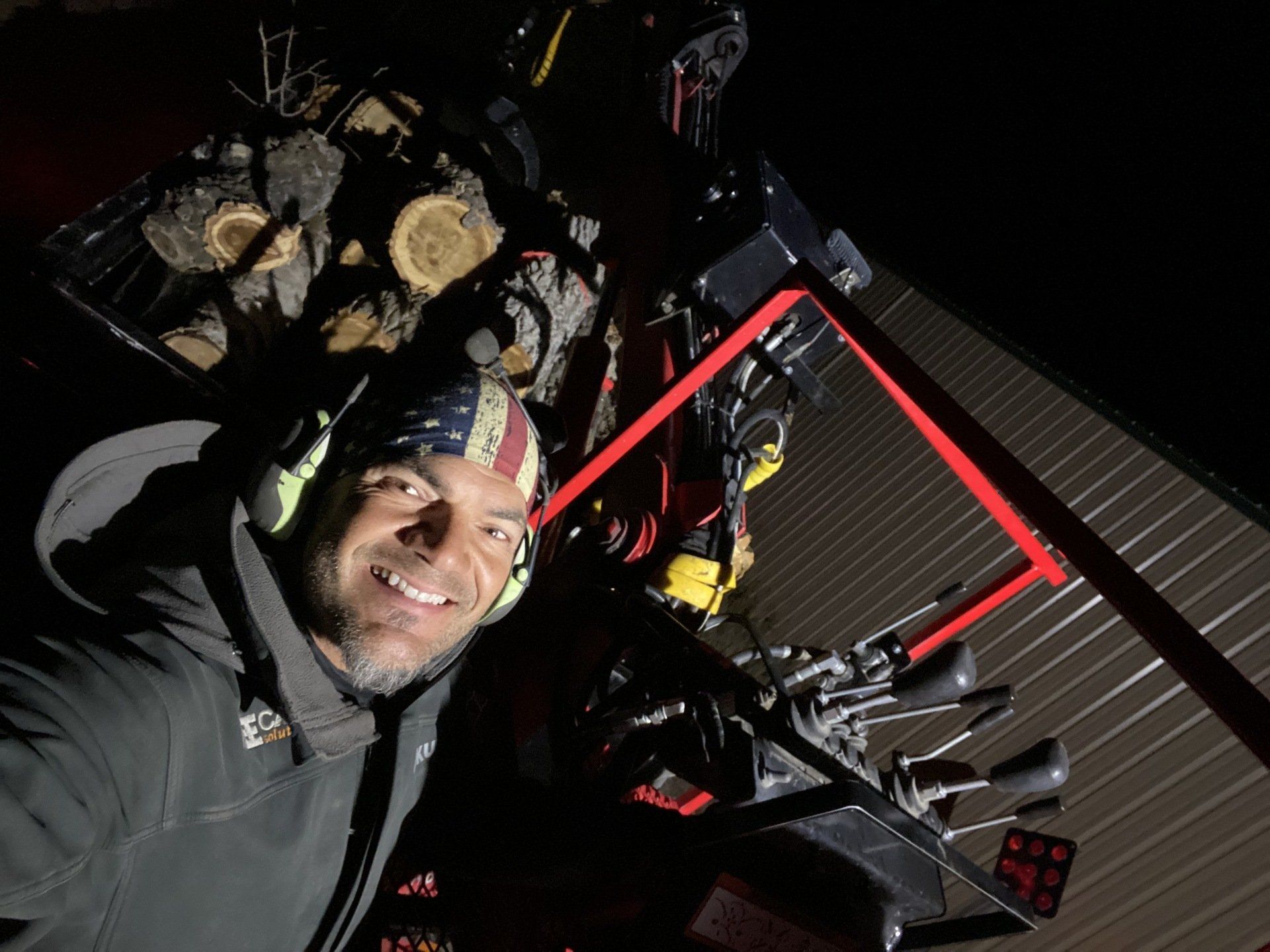 A man wearing an american flag bandana is smiling in front of a pile of logs.