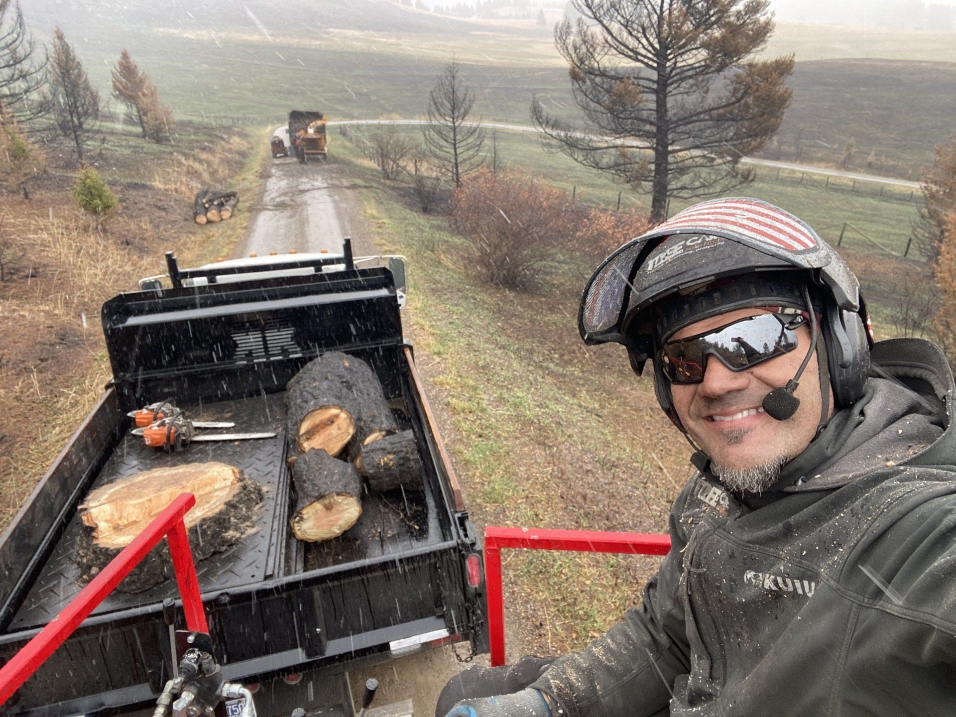 A man wearing a helmet and sunglasses is standing next to a trailer filled with logs.