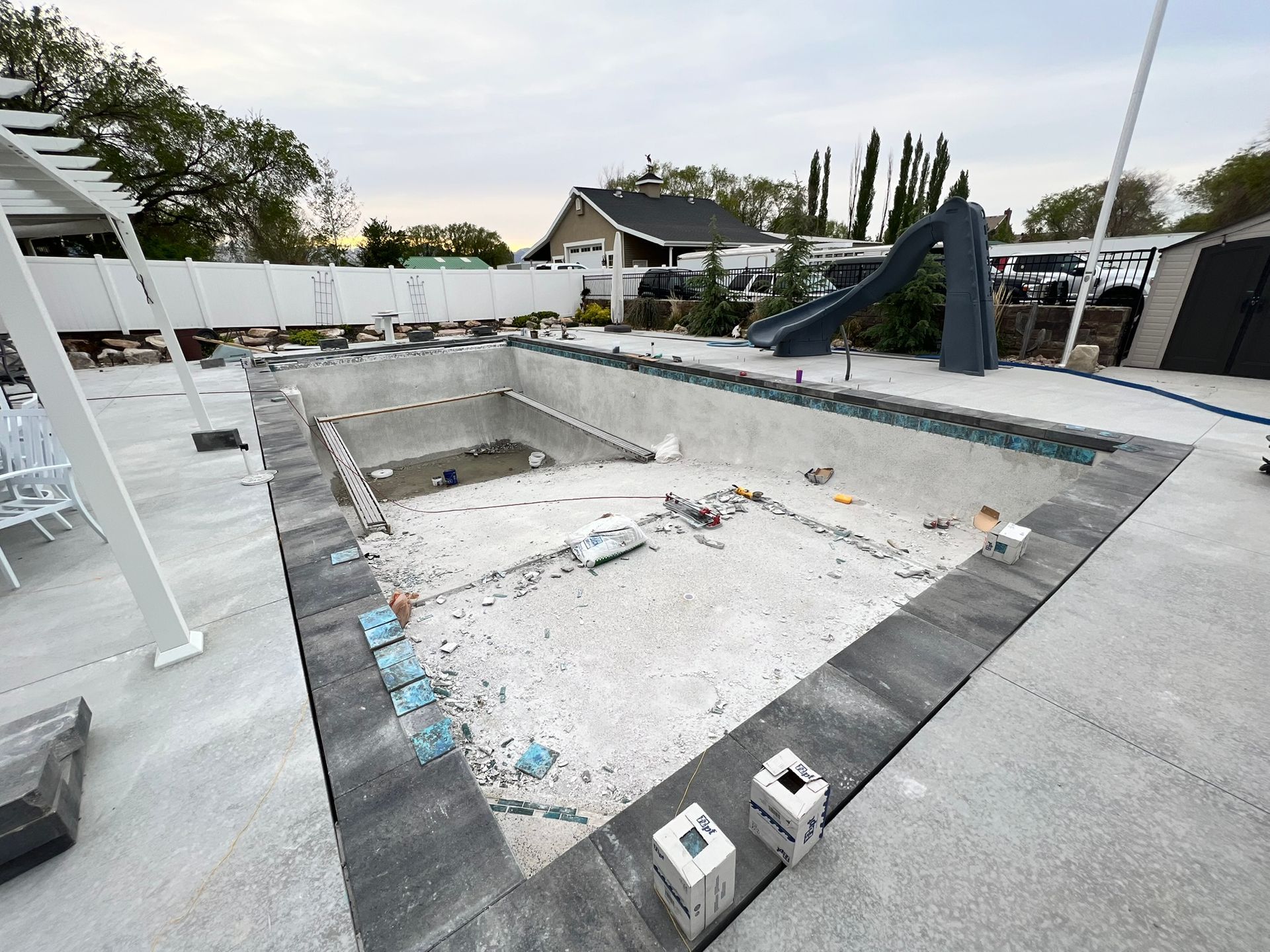 An empty swimming pool under construction, featuring gray stone coping, a slide, and a surrounding concrete deck.