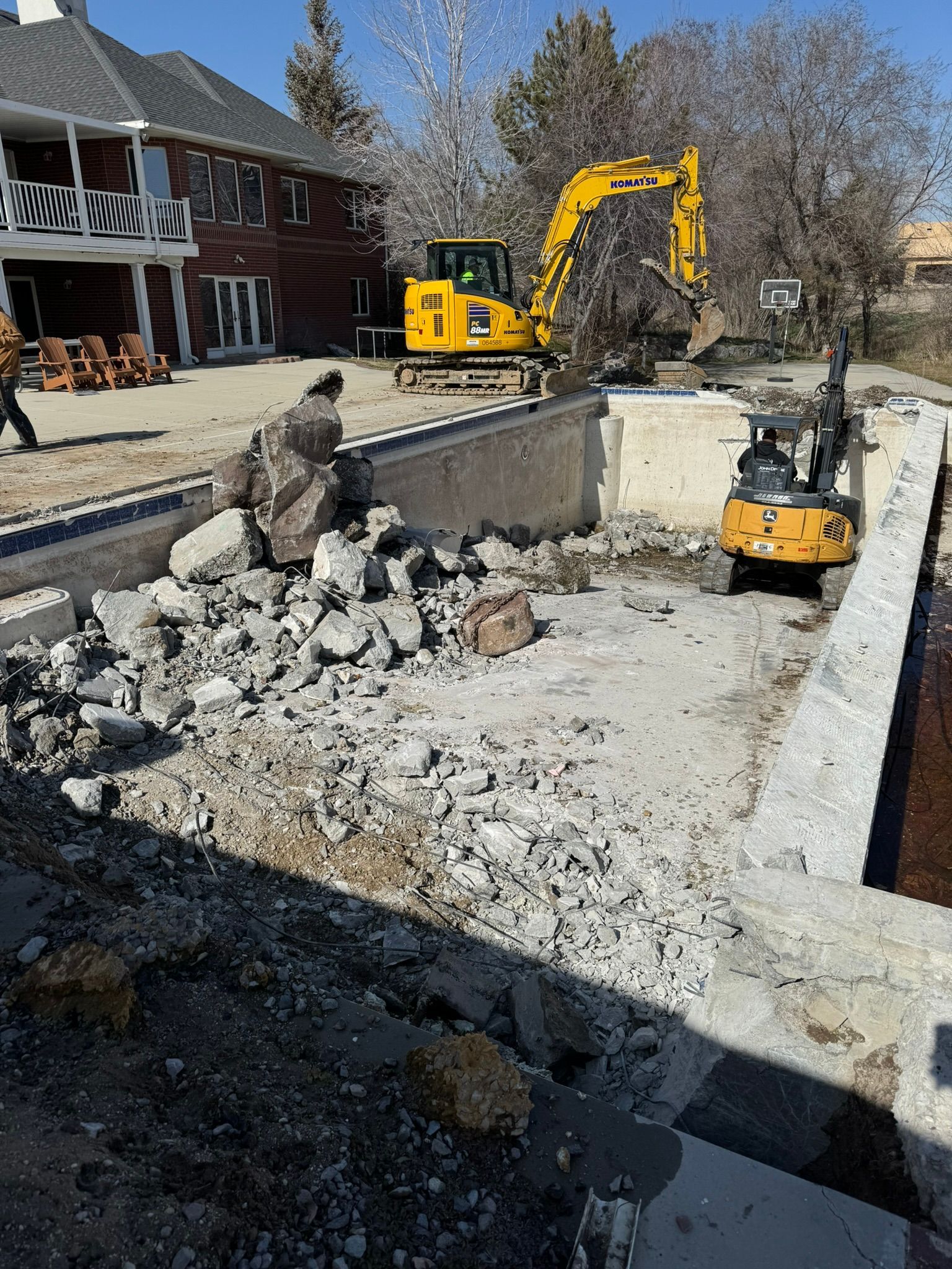 Construction crews use a yellow excavator to demolish an empty residential concrete swimming pool.
