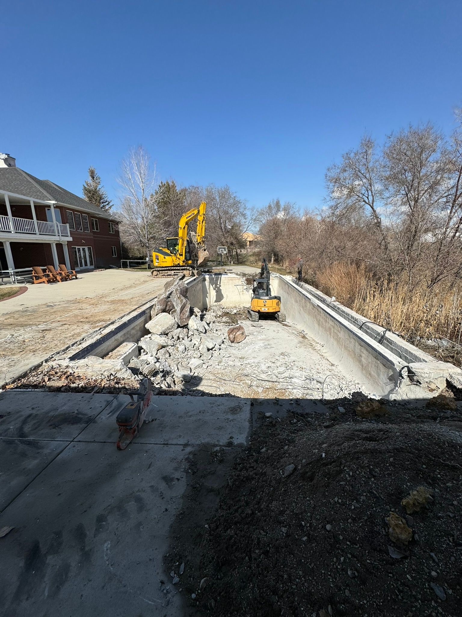 Yellow construction excavators demolish a concrete in-ground swimming pool behind a house on a sunny day.