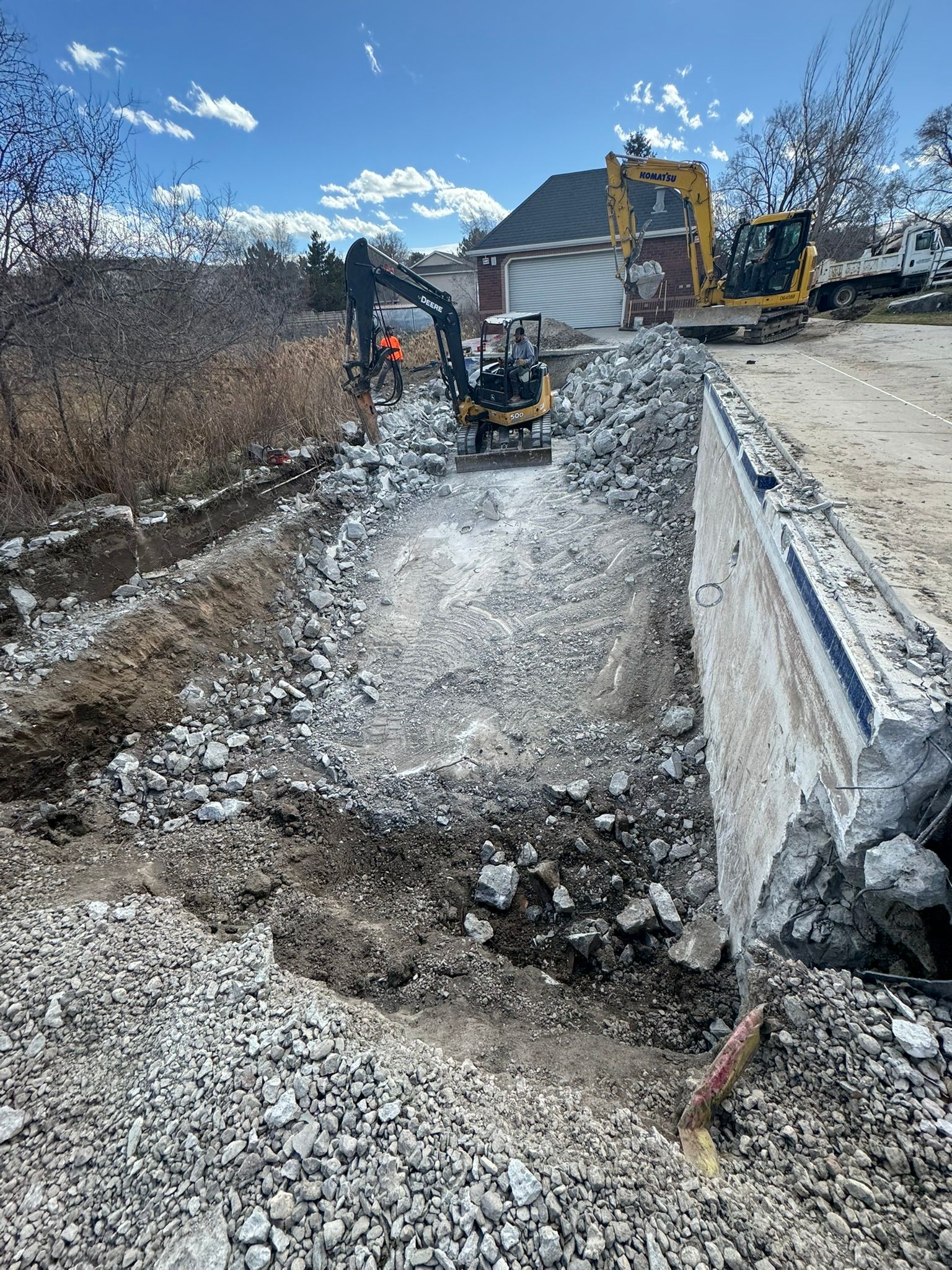 A small excavator works on a construction site, breaking up concrete rubble in an open trench near a large house.