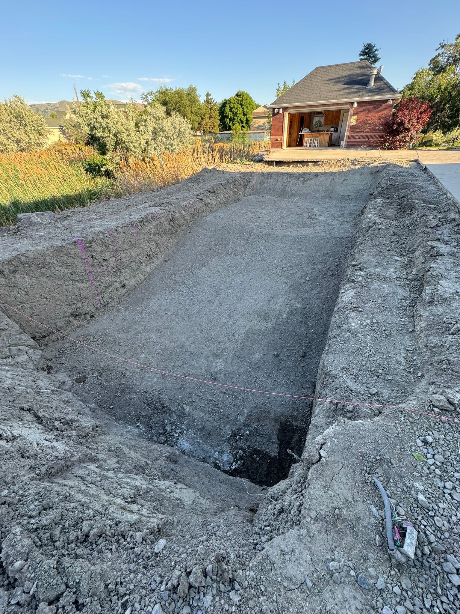 An excavated rectangular hole in a gravel-covered yard, with a house visible in the background.