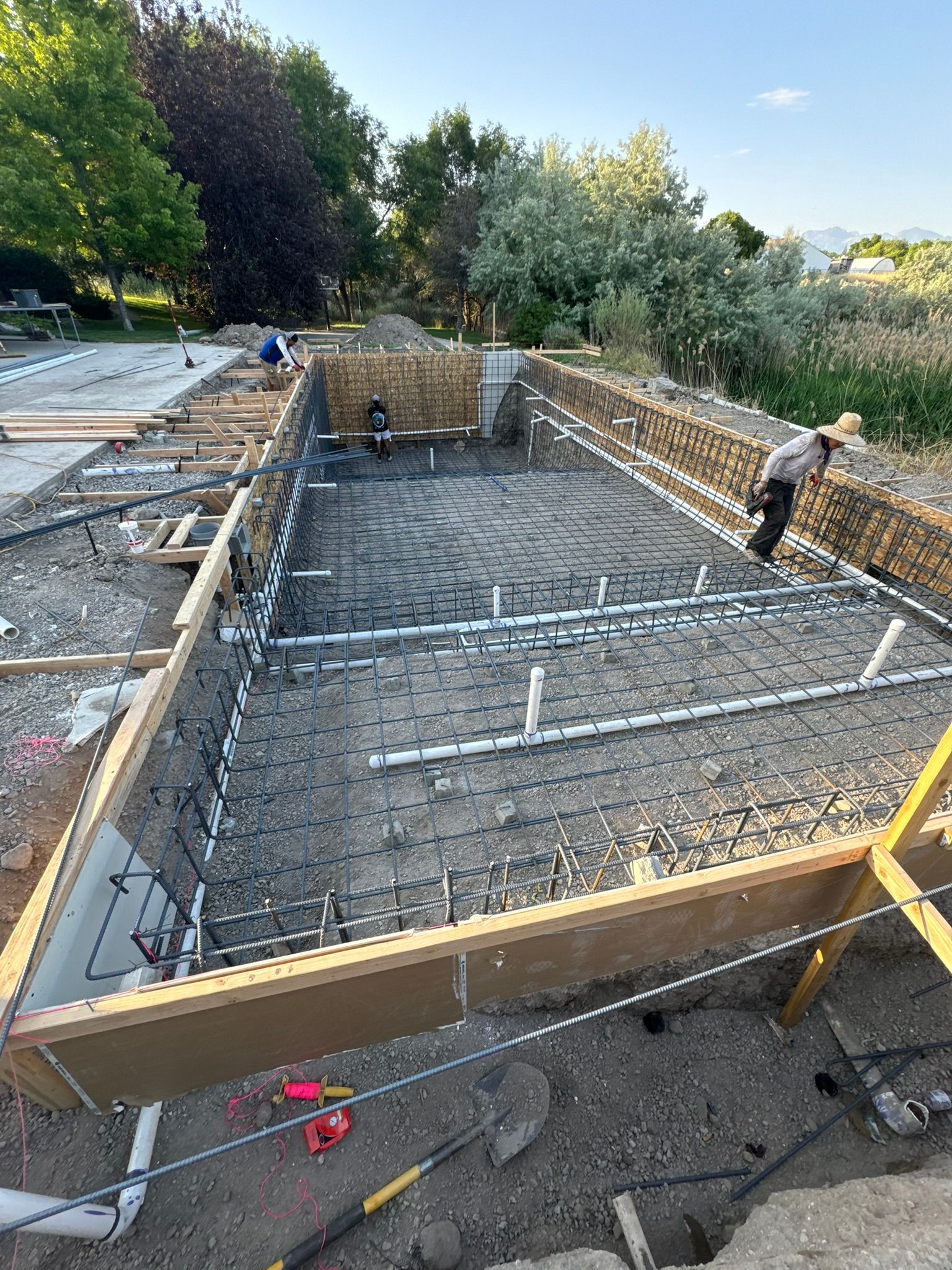 Workers installing plumbing and steel rebar framework in an excavated rectangular pool construction site.