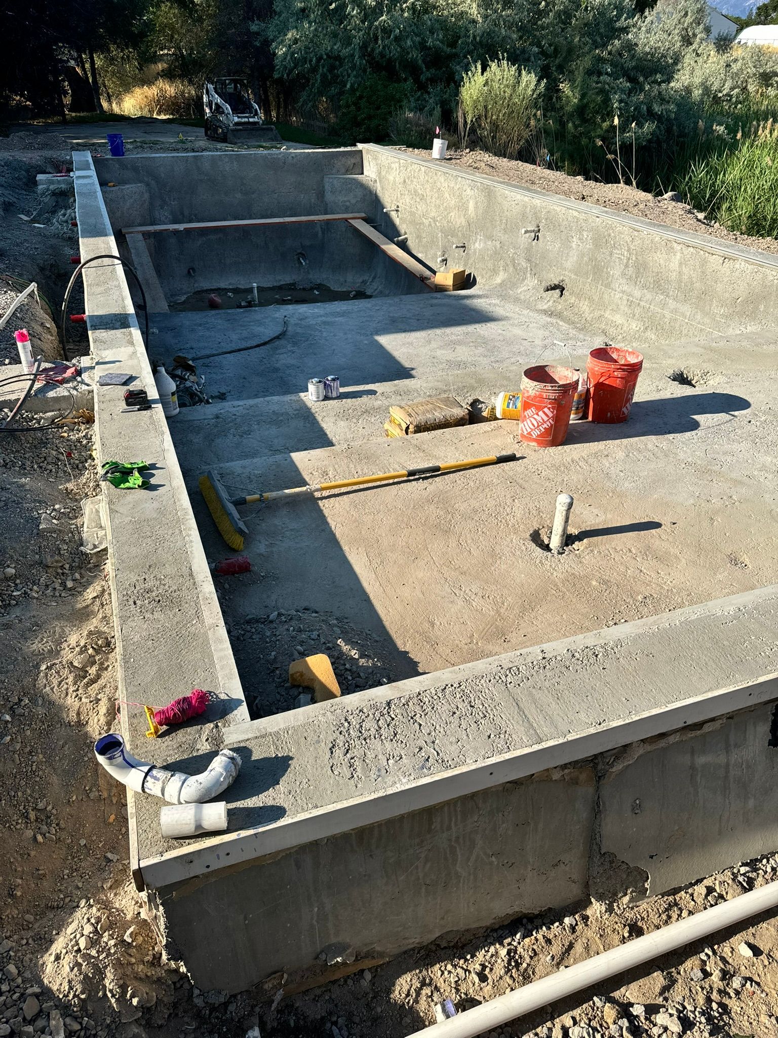 An unfinished concrete swimming pool structure with construction tools, buckets, and pipes on the ground.