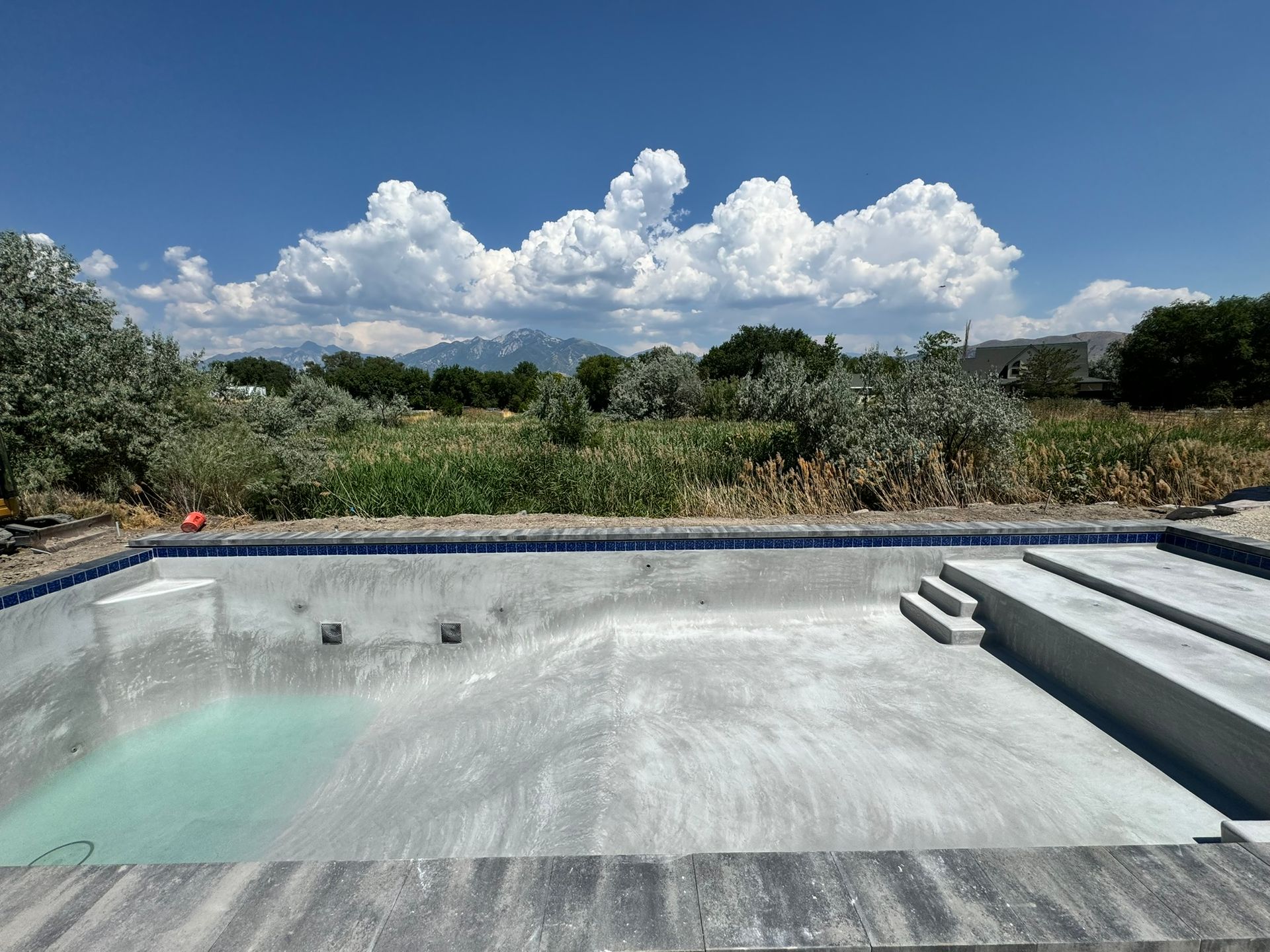 An empty, unfinished concrete swimming pool outdoors under a blue sky with fluffy white clouds and distant mountains.