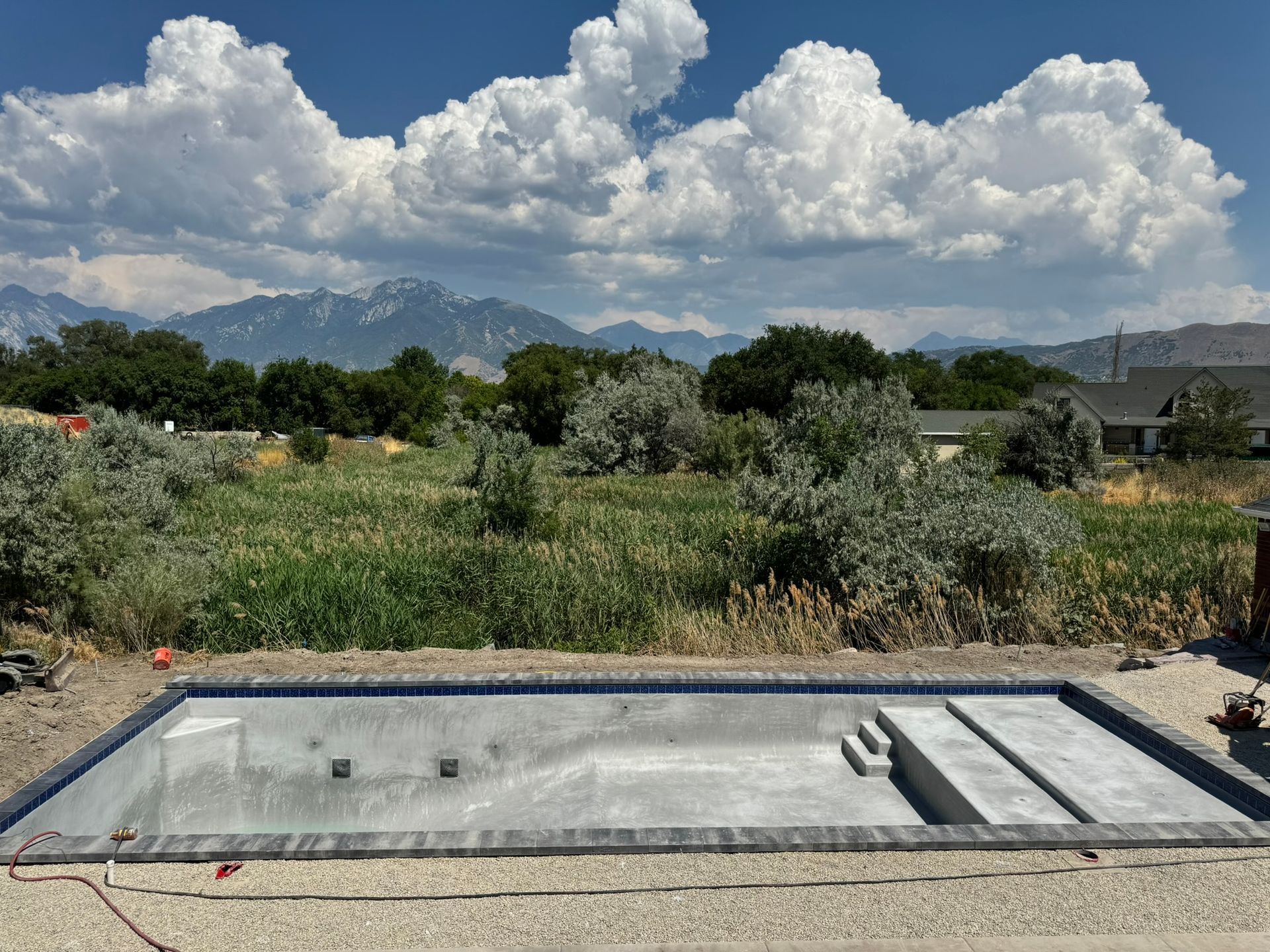 An unfinished concrete swimming pool in a backyard, set against a backdrop of green trees and distant mountains.
