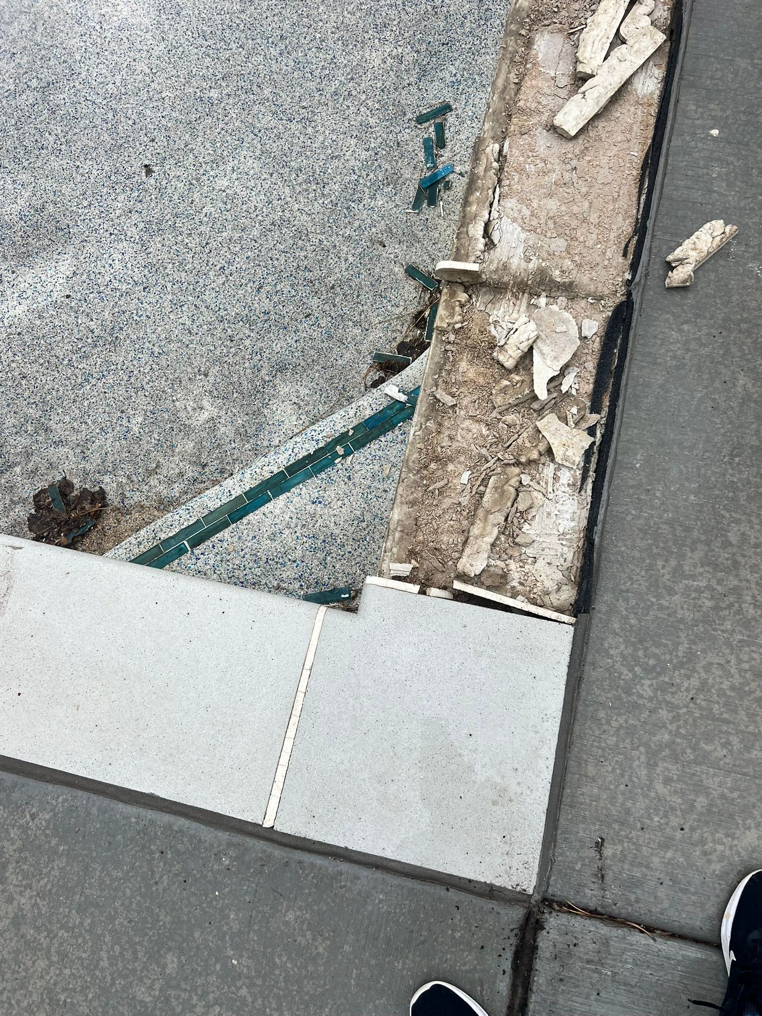 A section of a swimming pool edge showing missing coping tiles and exposed concrete, with adjacent pavers.