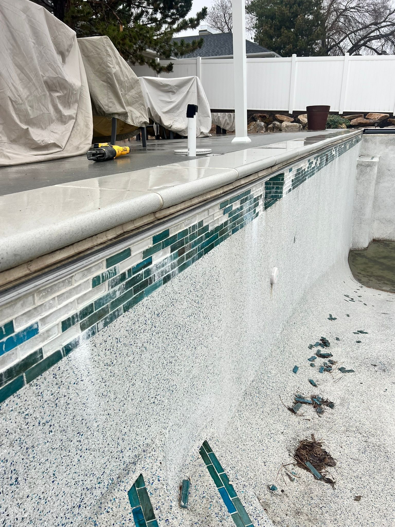 A close-up of a swimming pool edge showing damaged, detached blue mosaic tiles below the stone coping and speckled plaster.