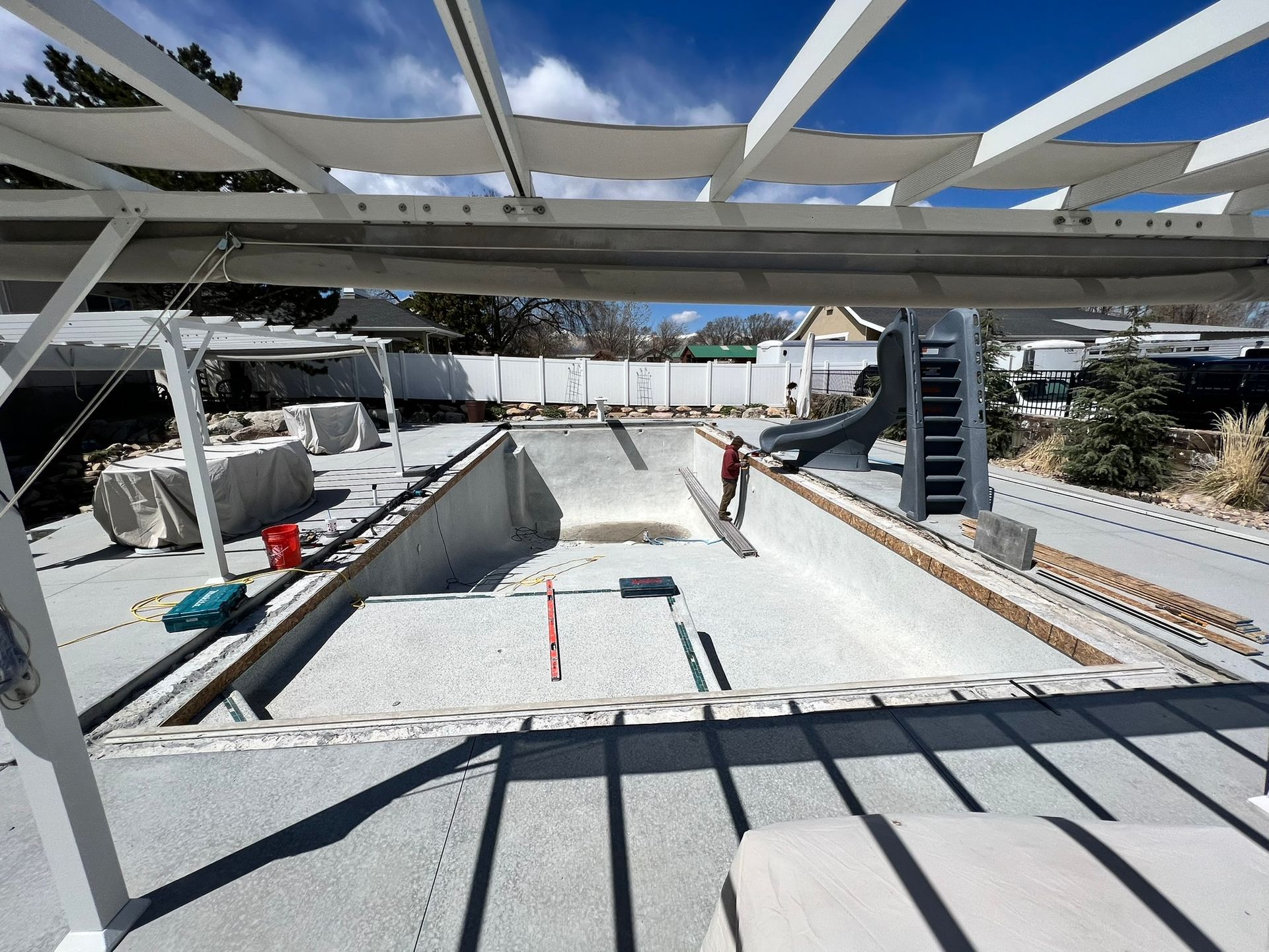 A person works inside an empty, rectangular pool surrounded by a patio and white pergola under a bright, sunny sky.