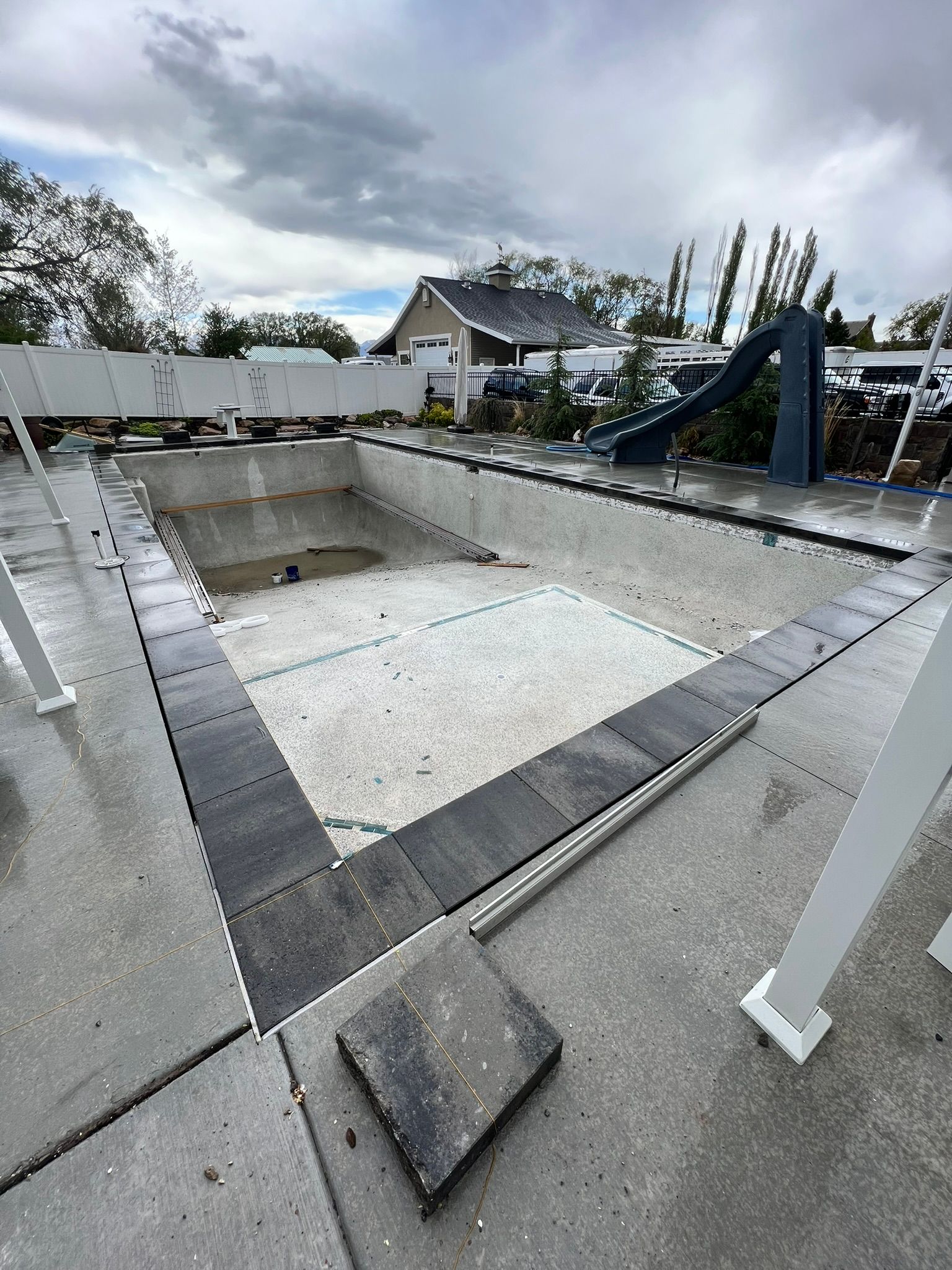 An empty backyard swimming pool under construction, featuring gray stone coping, concrete decking, and a blue slide.