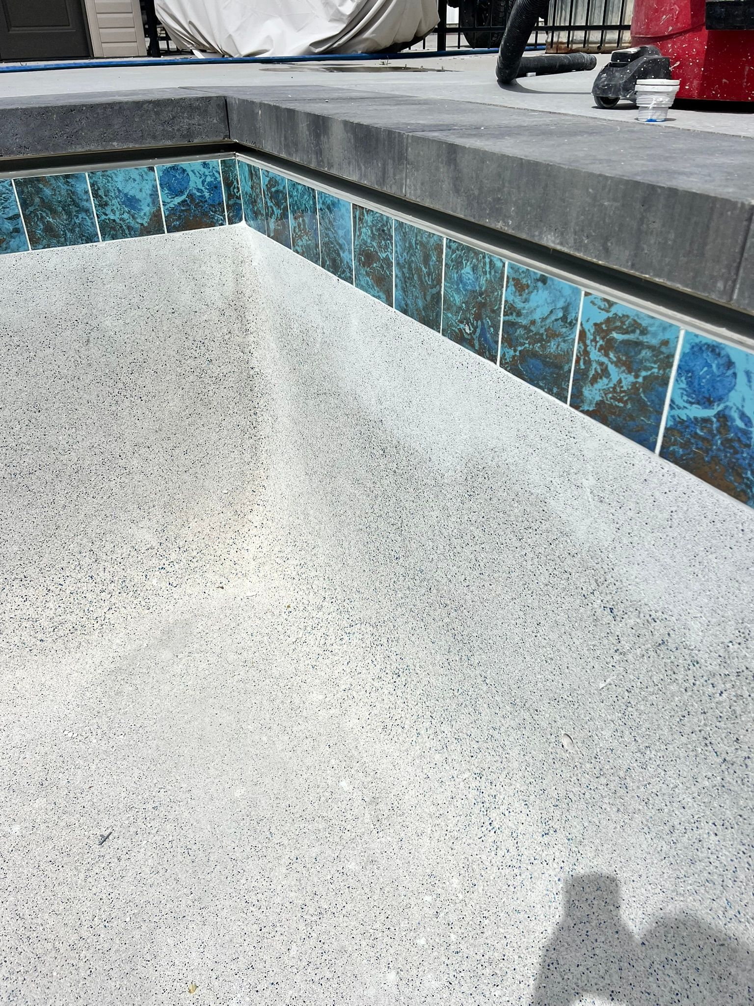 A corner of an empty swimming pool with light speckled plaster, blue tiled trim, and grey stone coping above.