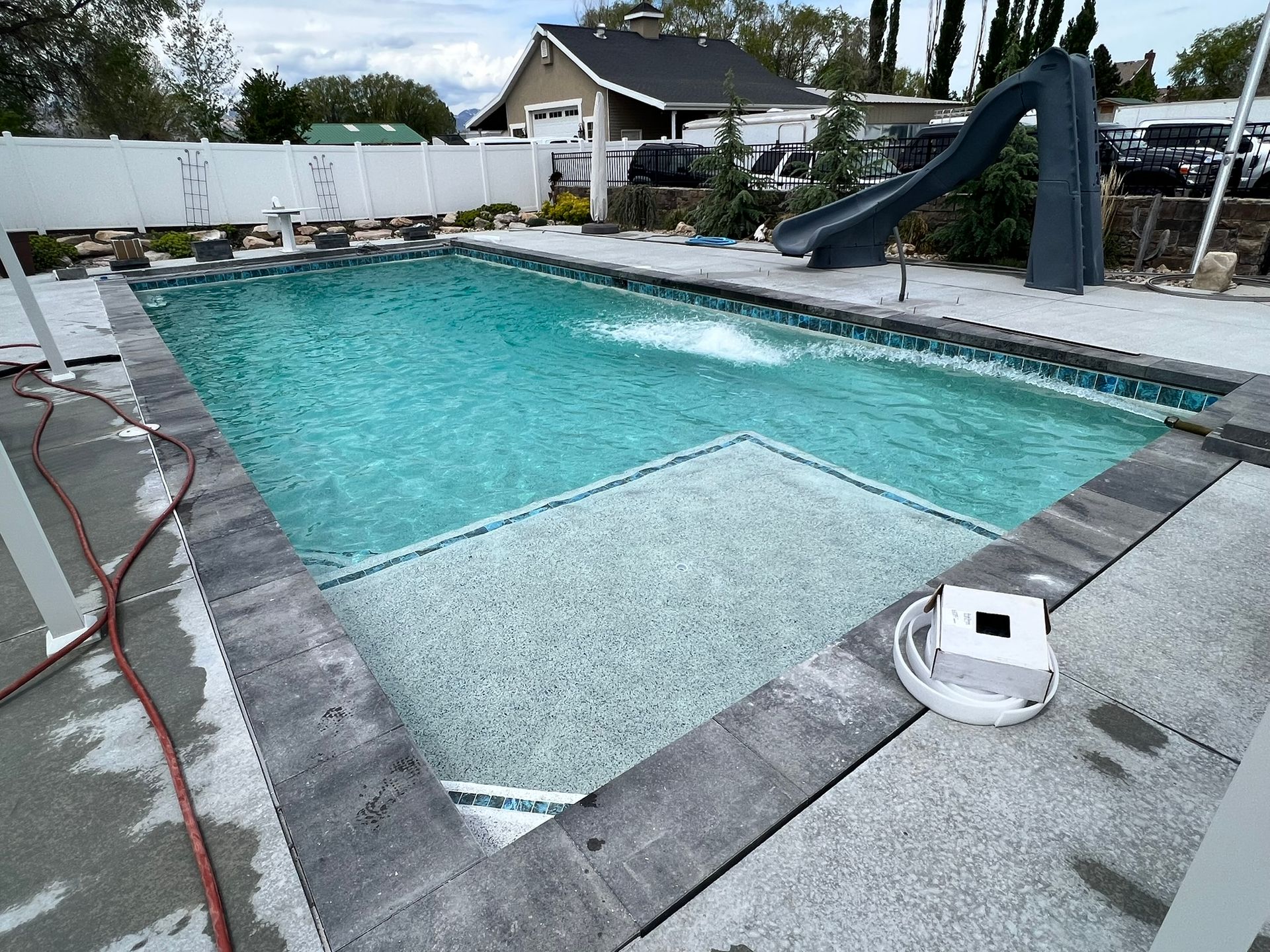 A rectangular backyard swimming pool with a shallow tanning ledge, surrounded by a dark stone border and white fence.