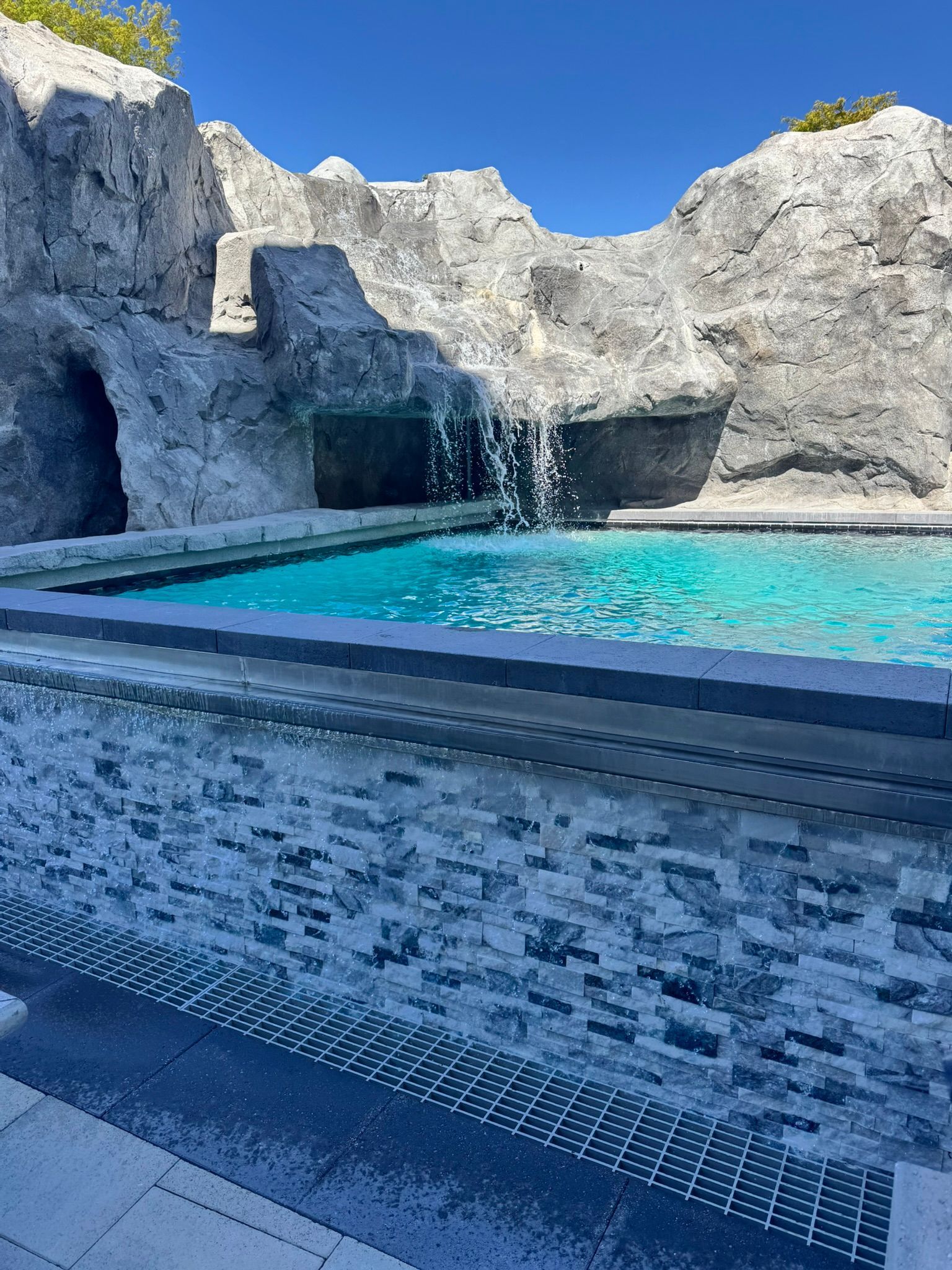 A swimming pool featuring a waterfall cascading from a large, rugged gray rock formation under a bright blue sky.