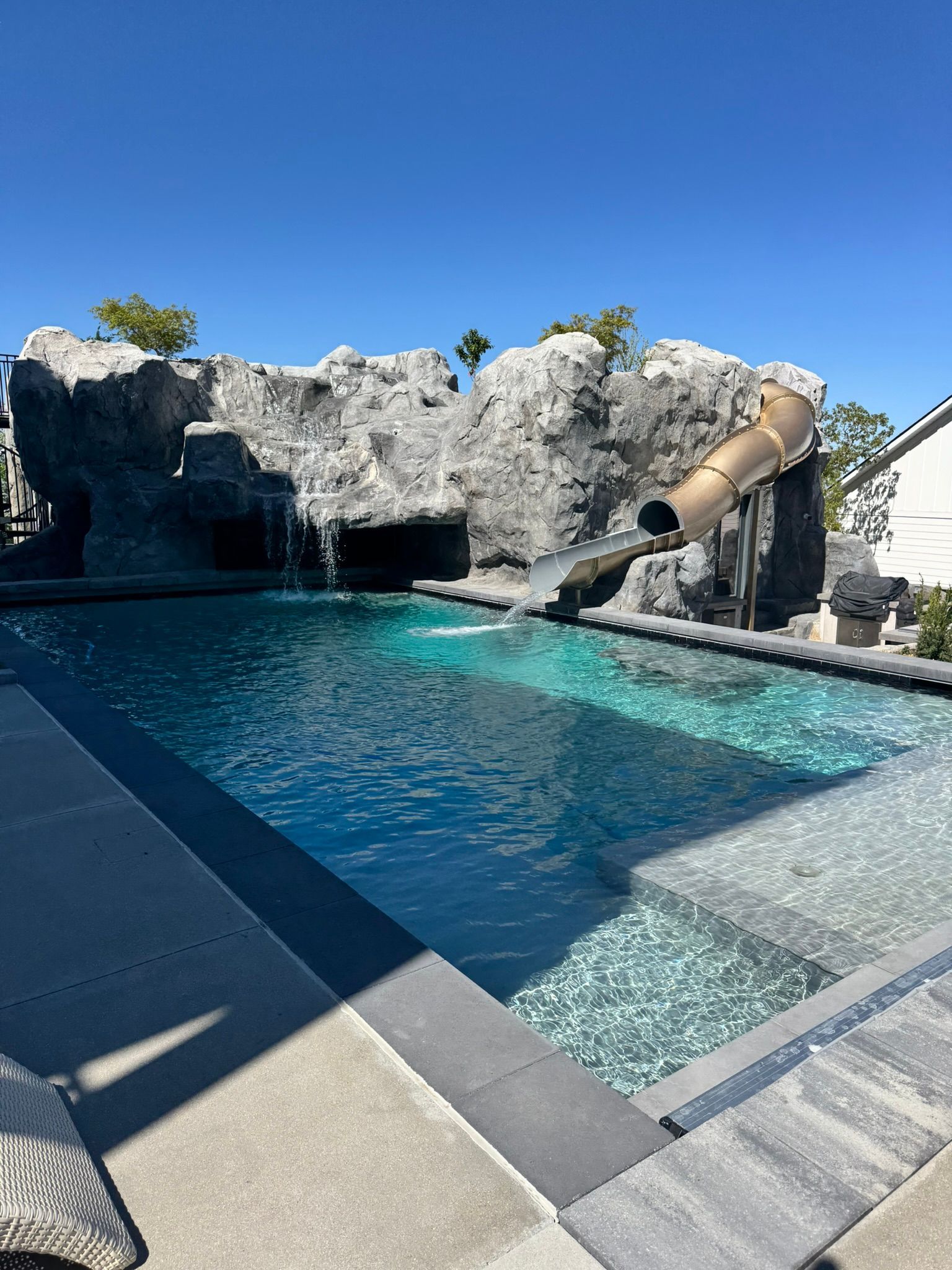 A backyard swimming pool with a large rock formation, waterfall, and a waterslide on a bright, sunny day.