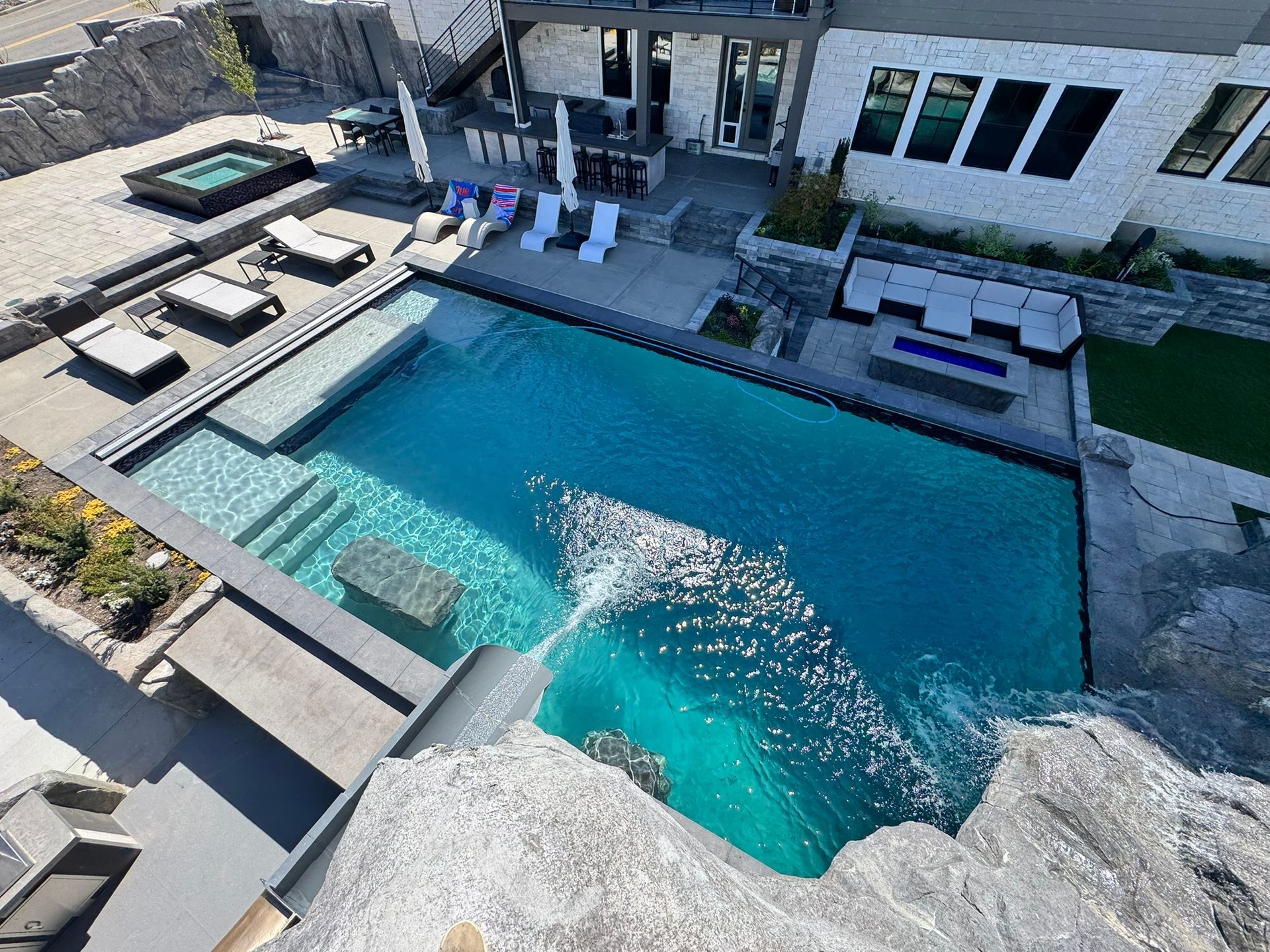 An aerial view of a rectangular backyard pool with a waterfall feature, lounge chairs, a hot tub, and a patio.